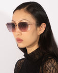 A person with long dark hair is wearing stylish Cece sunglasses from For Art's Sake®, featuring gradient lenses and a hexagonal frame. They are also wearing a black lace top and small earrings. The background is plain and neutral.