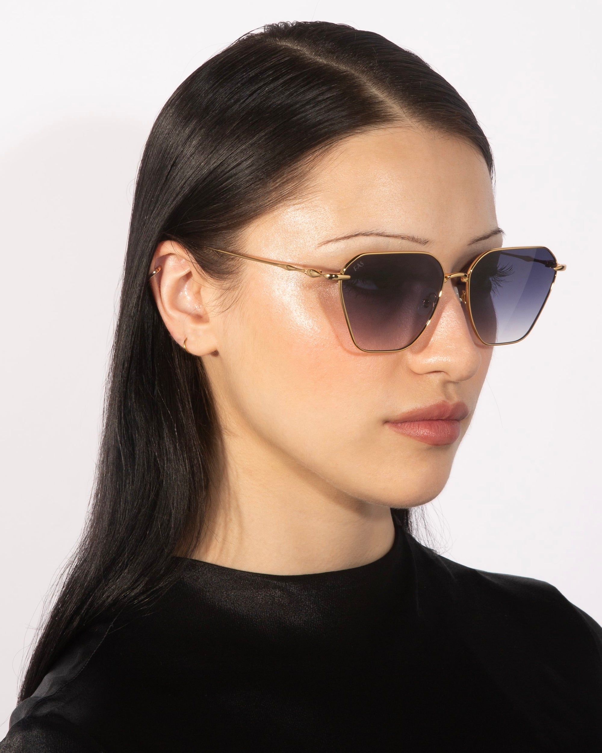 A person with long black hair is wearing stylish Cece sunglasses from For Art&#39;s Sake®, which feature a geometric shape, gold detailing, and gradient lenses. They are dressed in a black top and looking slightly to their left against a plain white background.