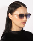 A person with long black hair is wearing stylish Cece sunglasses from For Art's Sake®, which feature a geometric shape, gold detailing, and gradient lenses. They are dressed in a black top and looking slightly to their left against a plain white background.