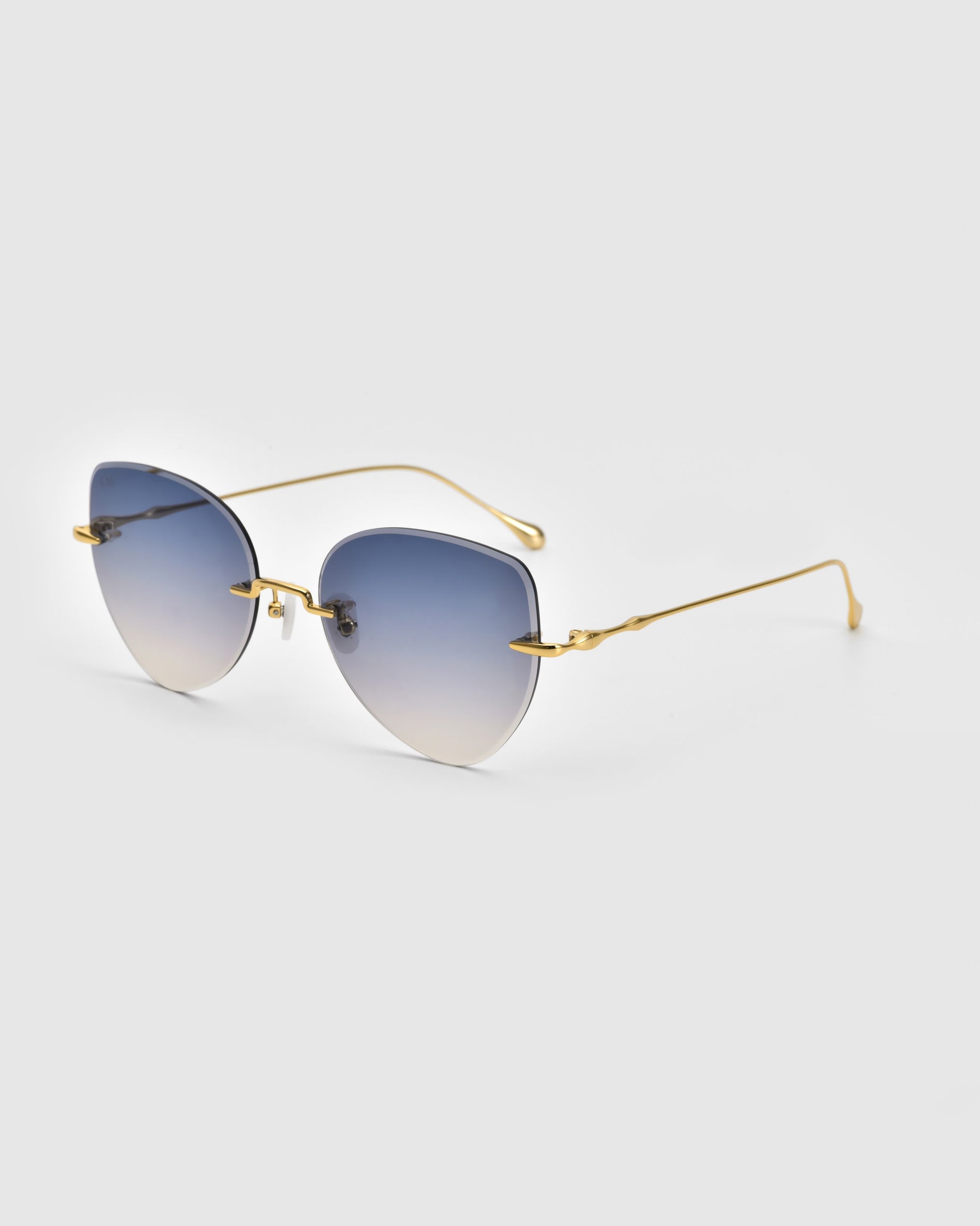 A pair of stylish Fifi Sunglasses by For Art&#39;s Sake®, featuring gradient blue lenses and thin 18k gold-plated frames. The design boasts sleek, minimalist temples and a cat-eye frameless silhouette, offering a modern and elegant look against a plain, light background.