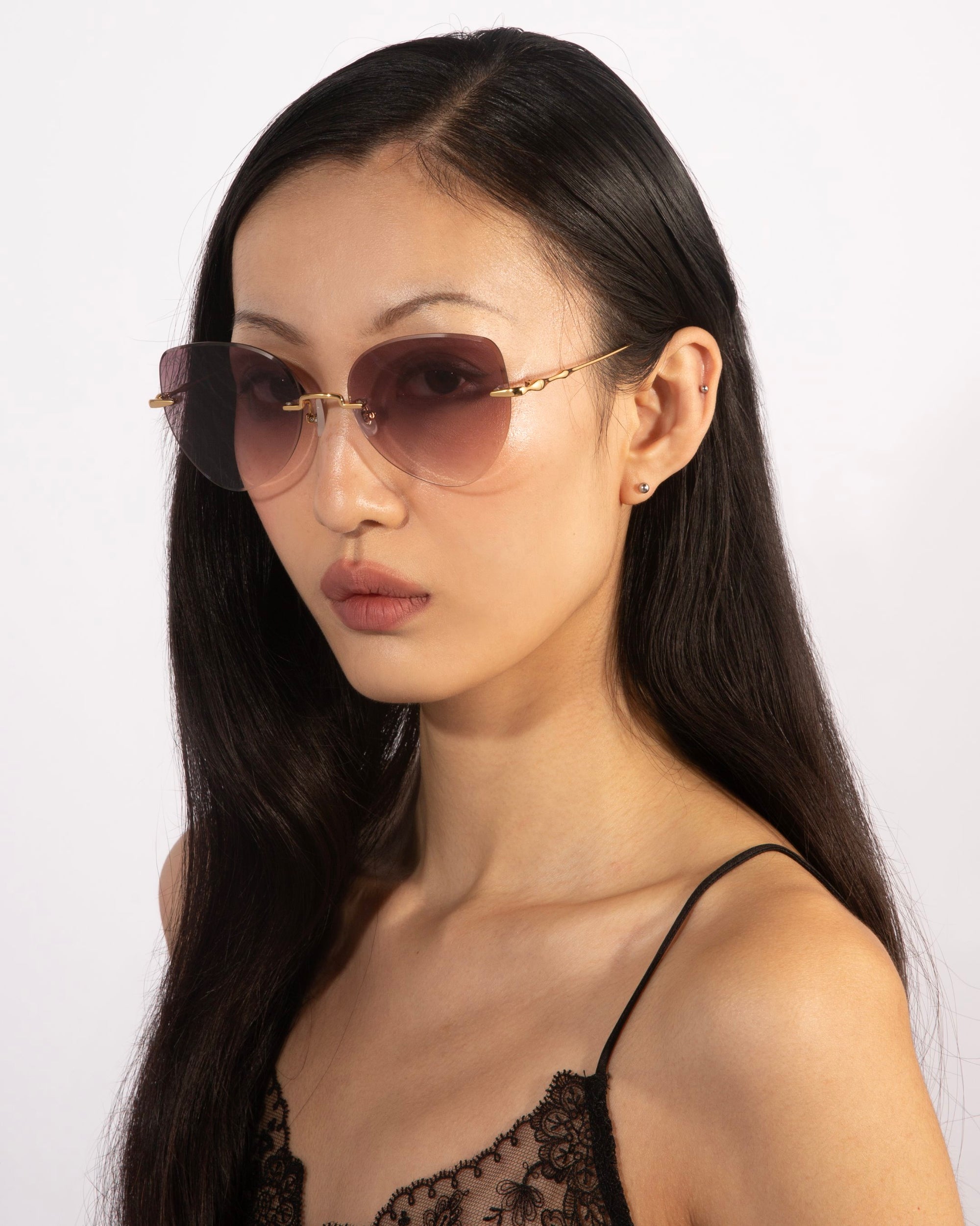 A person with long dark hair is wearing Fifi sunglasses from For Art&#39;s Sake®, featuring tinted lenses and thin frames. They are dressed in a black lace strap top and looking into the camera against a plain white background.