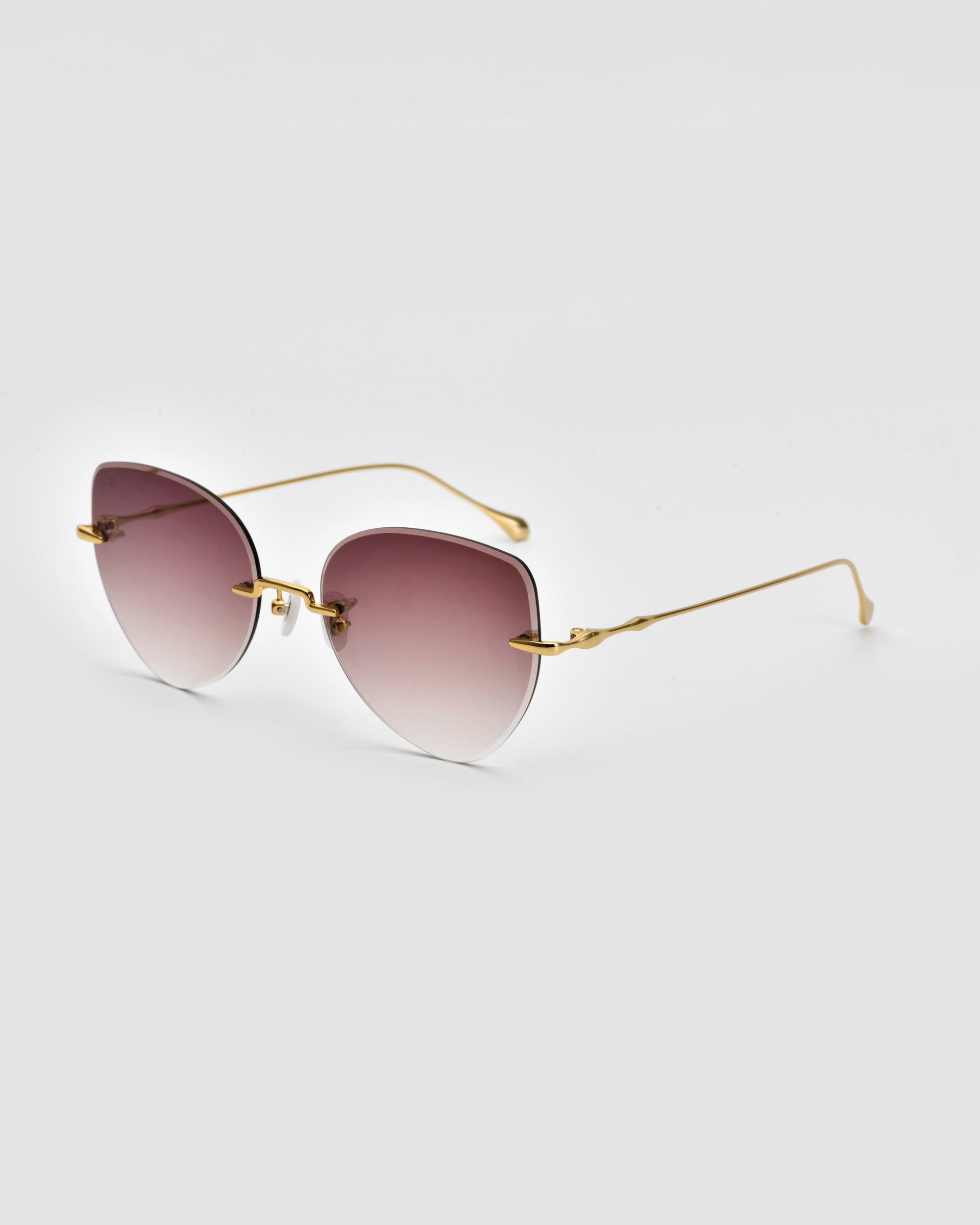 Introducing the Fifi Sunglasses by For Art&#39;s Sake®: a chic pair of stylish cat-eye frameless sunglasses featuring gradient lenses that transition from dark at the top to lighter at the bottom, complemented by gold-colored thin metal frames and arms. The backdrop is a plain white.