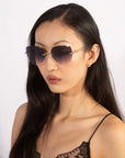 A woman with long, straight black hair is wearing Fifi sunglasses from For Art's Sake®, which feature gradient lenses and a gold frame with thin temples. She has a neutral facial expression and is dressed in a black spaghetti-strap top with lace detailing. The background is white and plain.