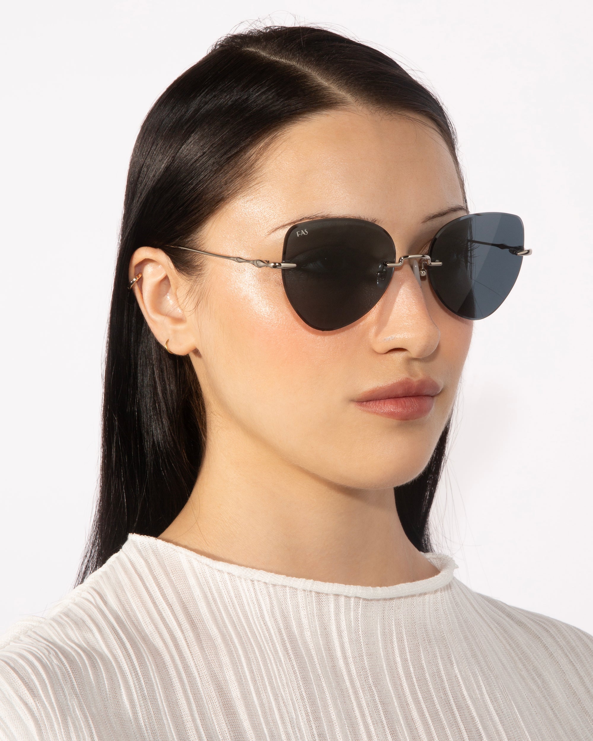 A woman with long, dark hair sports the Fifi sunglasses by For Art's Sake®, featuring dark lenses and a cat-eye shape. She has a neutral expression and is dressed in a white ribbed top. The background is plain and white.