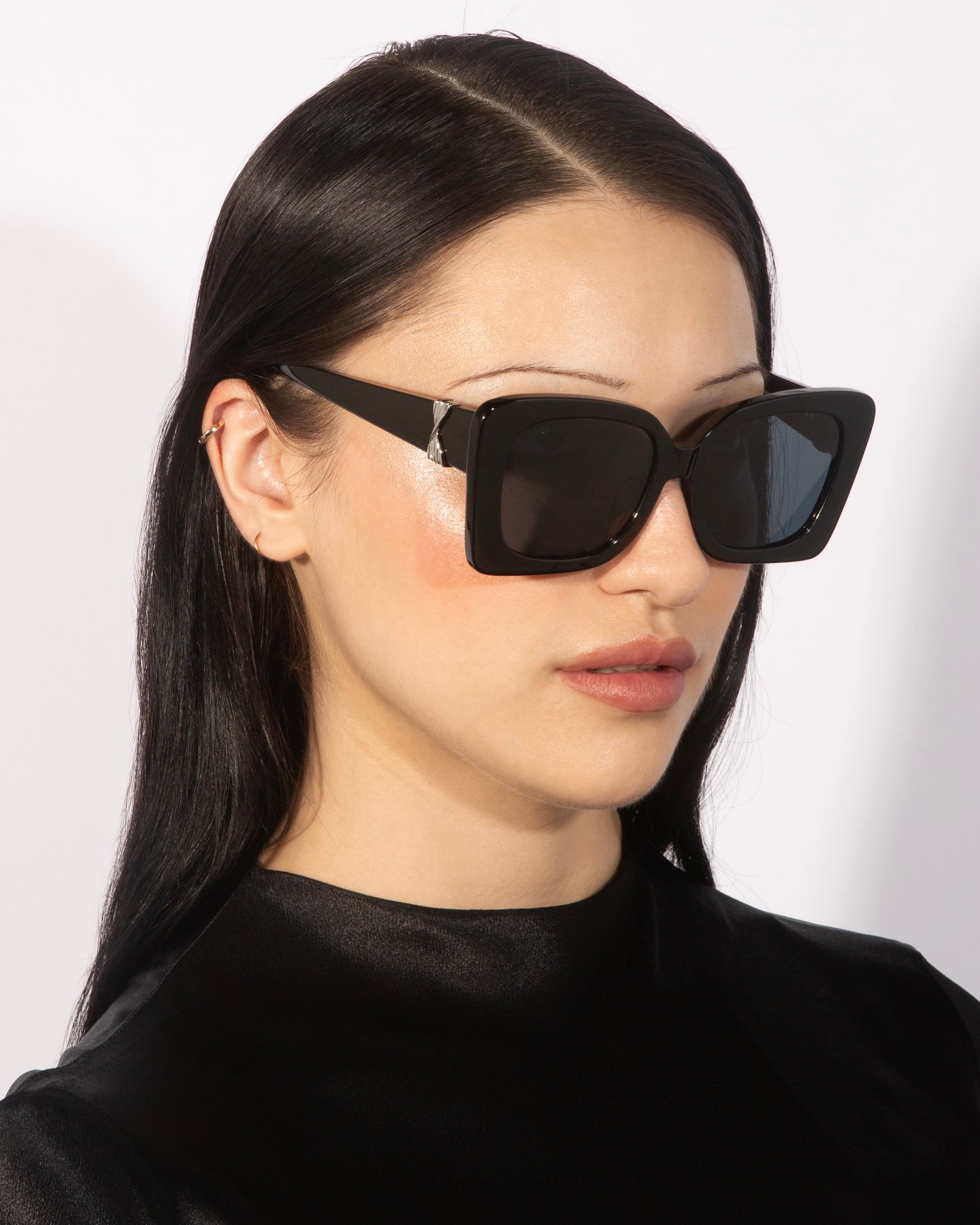 A person with long, straight black hair is wearing the &quot;Ribbon&quot; oversized square cat-eye sunglasses by For Art&#39;s Sake® and a black, shiny, high-neck top. The background is simple and white, and the person has a neutral expression.