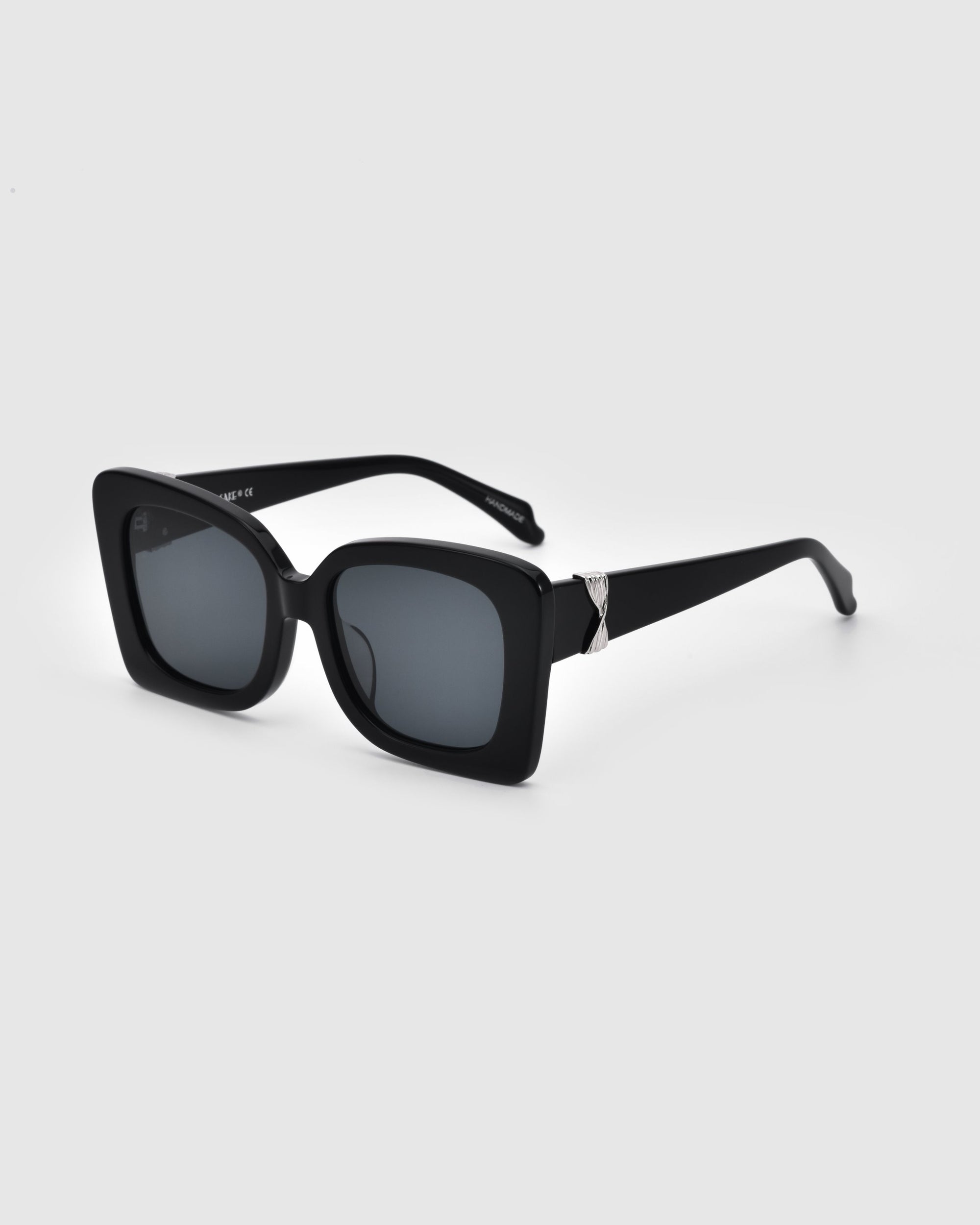 The Rectangular black Ribbon sunglasses from For Art&#39;s Sake® boast dark lenses and a thick, black frame. This chic accessory is enhanced with a silver decorative element near the hinges. The sustainable sunglasses are positioned at an angle to the left against a gray background.