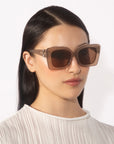 A person wearing the For Art's Sake® Ribbon oversized square brown sunglasses and a white textured top. They have straight, long, dark hair and a neutral expression, facing slightly to the left against a plain white background.