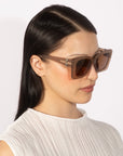 A woman with long, dark hair is wearing a white, textured sleeveless top and the Ribbon oversized square cat-eye sunglasses by For Art's Sake®. The background is plain and white, highlighting her sustainable luxury eyewear and attire. She is facing slightly to the side with a calm expression.