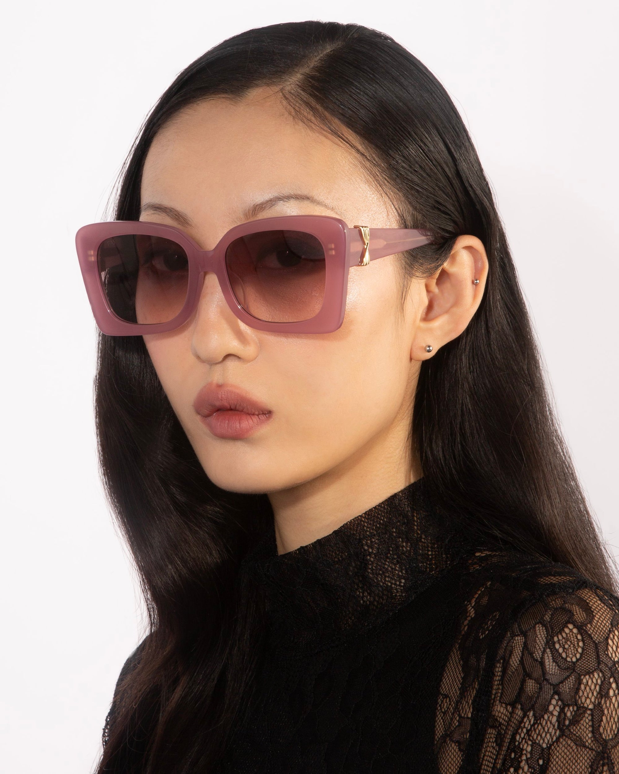 A person with long, dark hair is wearing oversized Ribbon sunglasses in pink by For Art&#39;s Sake® and a black lace top. They have a neutral facial expression and are positioned against a plain white background.