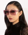 A person with long, dark hair is wearing oversized Ribbon sunglasses in pink by For Art's Sake® and a black lace top. They have a neutral facial expression and are positioned against a plain white background.