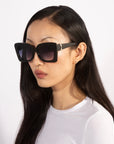 A person with long, straight black hair is wearing the Ribbon oversized, square black sunglasses from For Art's Sake®—a stylish accessory—and a white top. Their expression is neutral, and they are set against a plain white background.