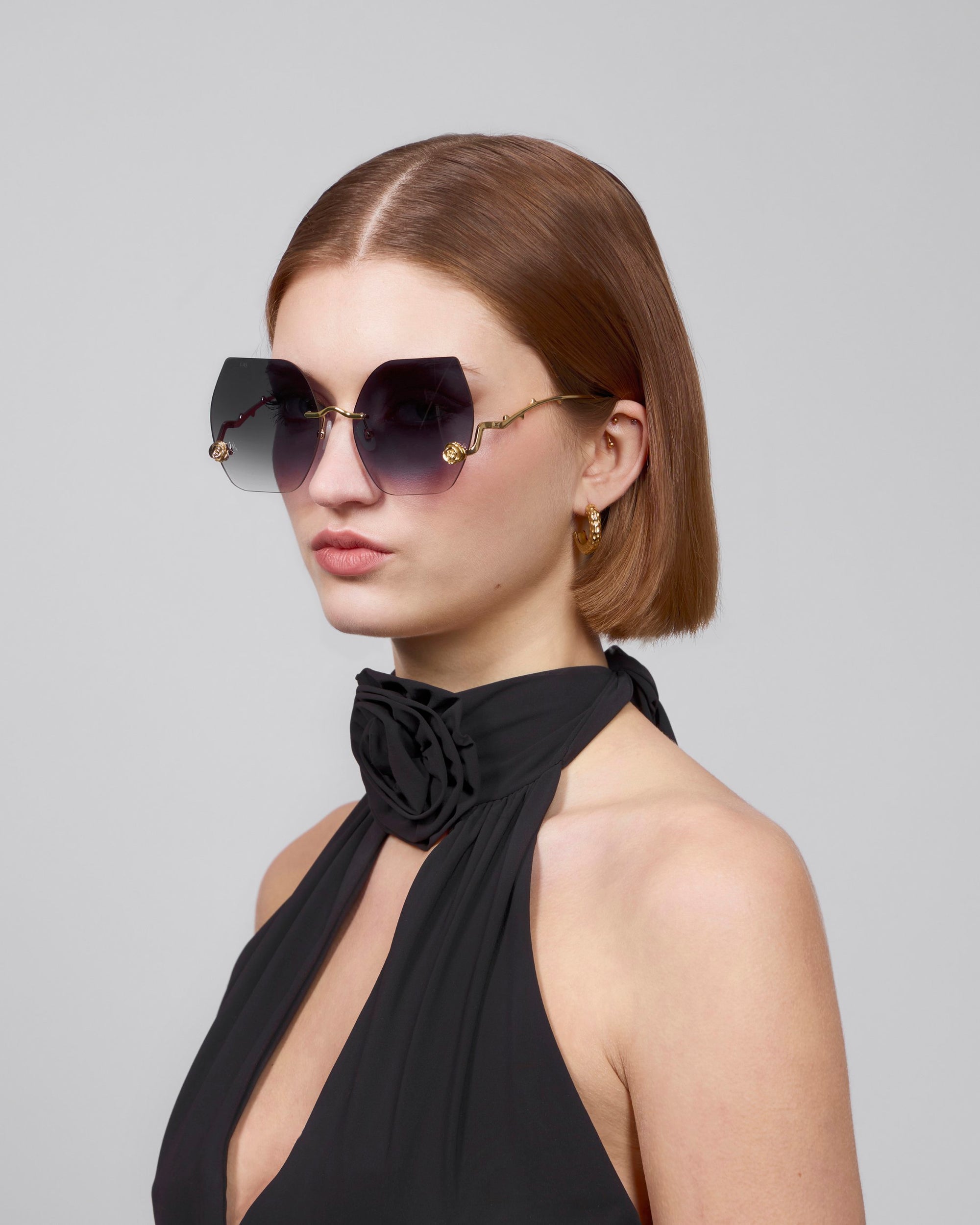 A woman with short auburn hair wears La Vie En Rose oversized square sunglasses made of biodegradable acetate by For Art's Sake®. She's in a black halter-neck top with a large floral detail, gold hoop earrings swaying as she gazes sideways against a plain background.