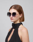 A woman with short auburn hair wears La Vie En Rose oversized square sunglasses made of biodegradable acetate by For Art's Sake®. She's in a black halter-neck top with a large floral detail, gold hoop earrings swaying as she gazes sideways against a plain background.