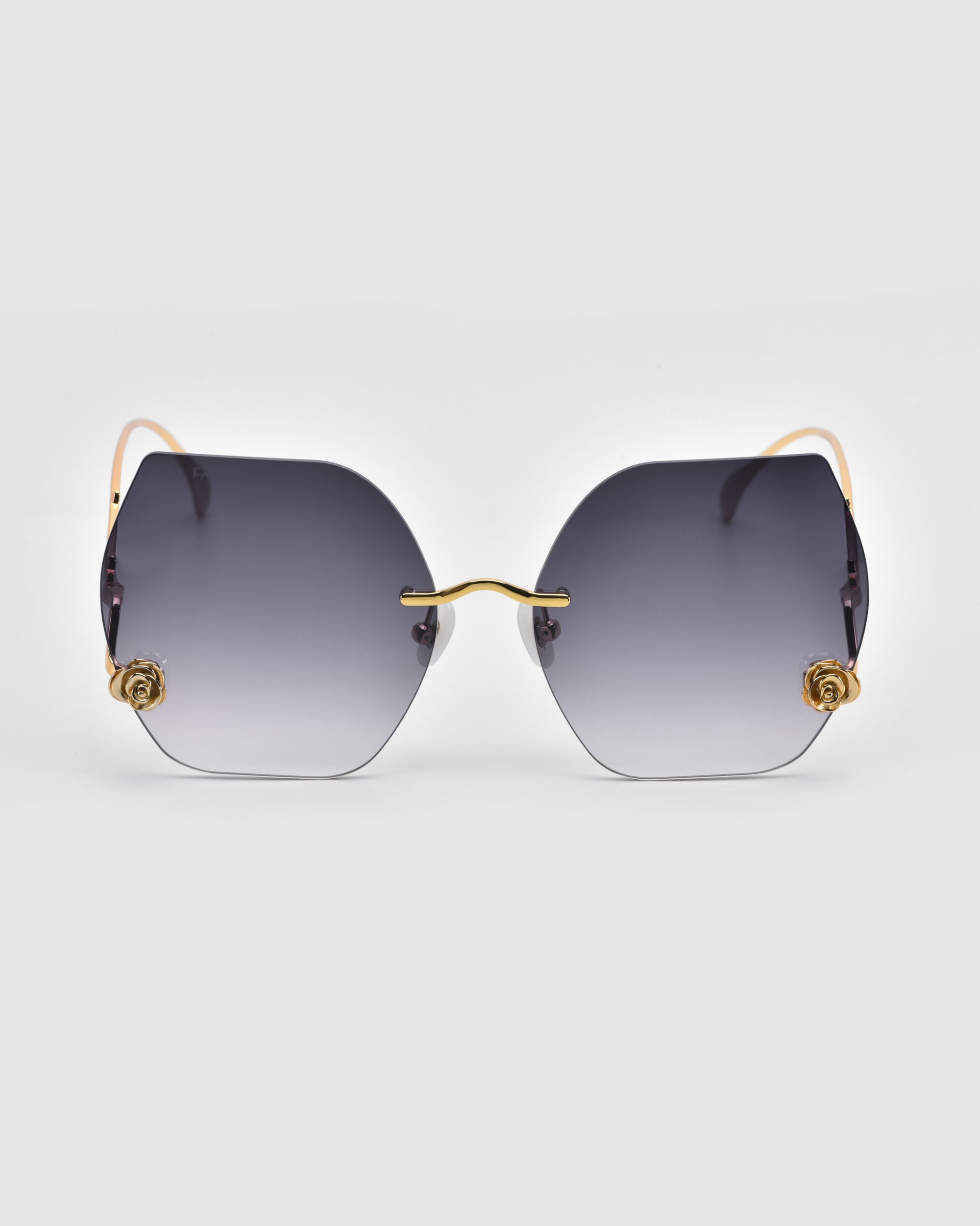 From For Art's Sake®'s La Vie En Rose collection, these hexagonal sunglasses feature gradient gray lenses and thin gold frames with rose details at the temples, offering sophistication on a white backdrop.