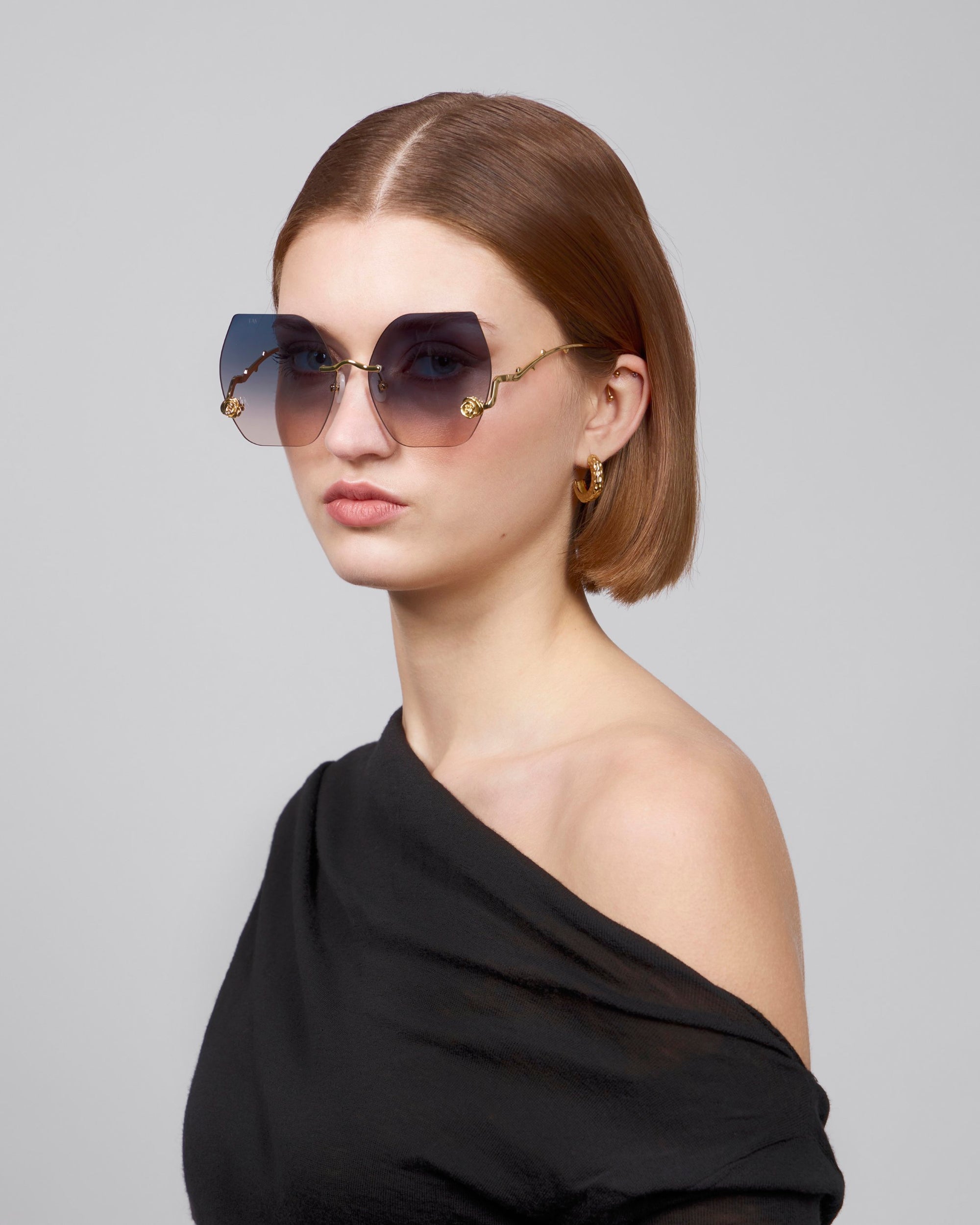 A woman donning oversized, square-shaped sunglasses crafted from retro-inspired biodegradable acetate by For Art's Sake® (La Vie En Rose) pairs them with gold hoop earrings and an off-the-shoulder black top, standing against a gray background. Her brown hair is shoulder-length.