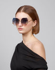 A woman donning oversized, square-shaped sunglasses crafted from retro-inspired biodegradable acetate by For Art's Sake® (La Vie En Rose) pairs them with gold hoop earrings and an off-the-shoulder black top, standing against a gray background. Her brown hair is shoulder-length.