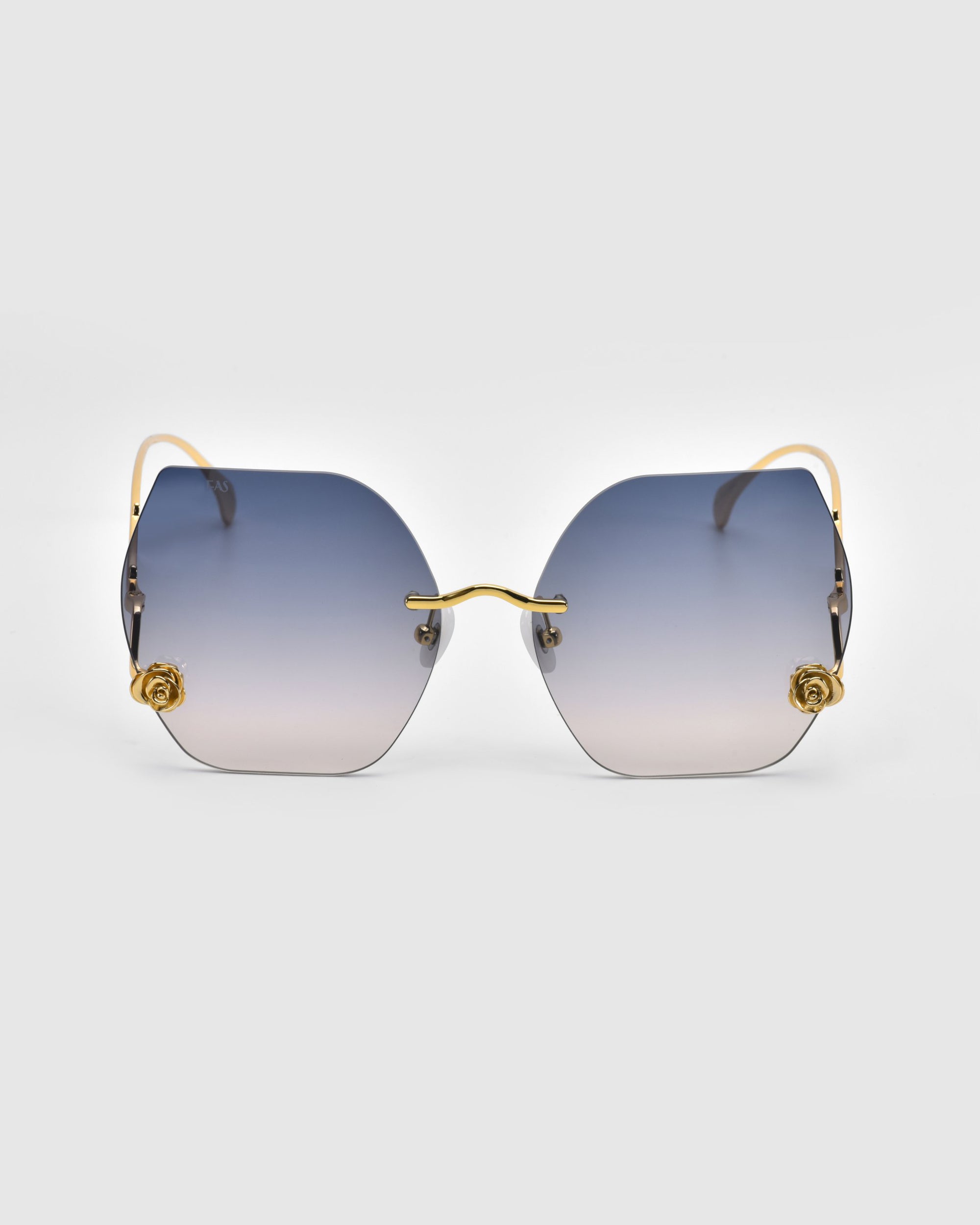 From the Rose Garden Collection, For Art's Sake® presents the La Vie En Rose hexagonal sunglasses with oversized frameless lenses that transition from blue to clear, featuring gold rose temple accents and slender gold arms against a soft gray background for an elegant look.