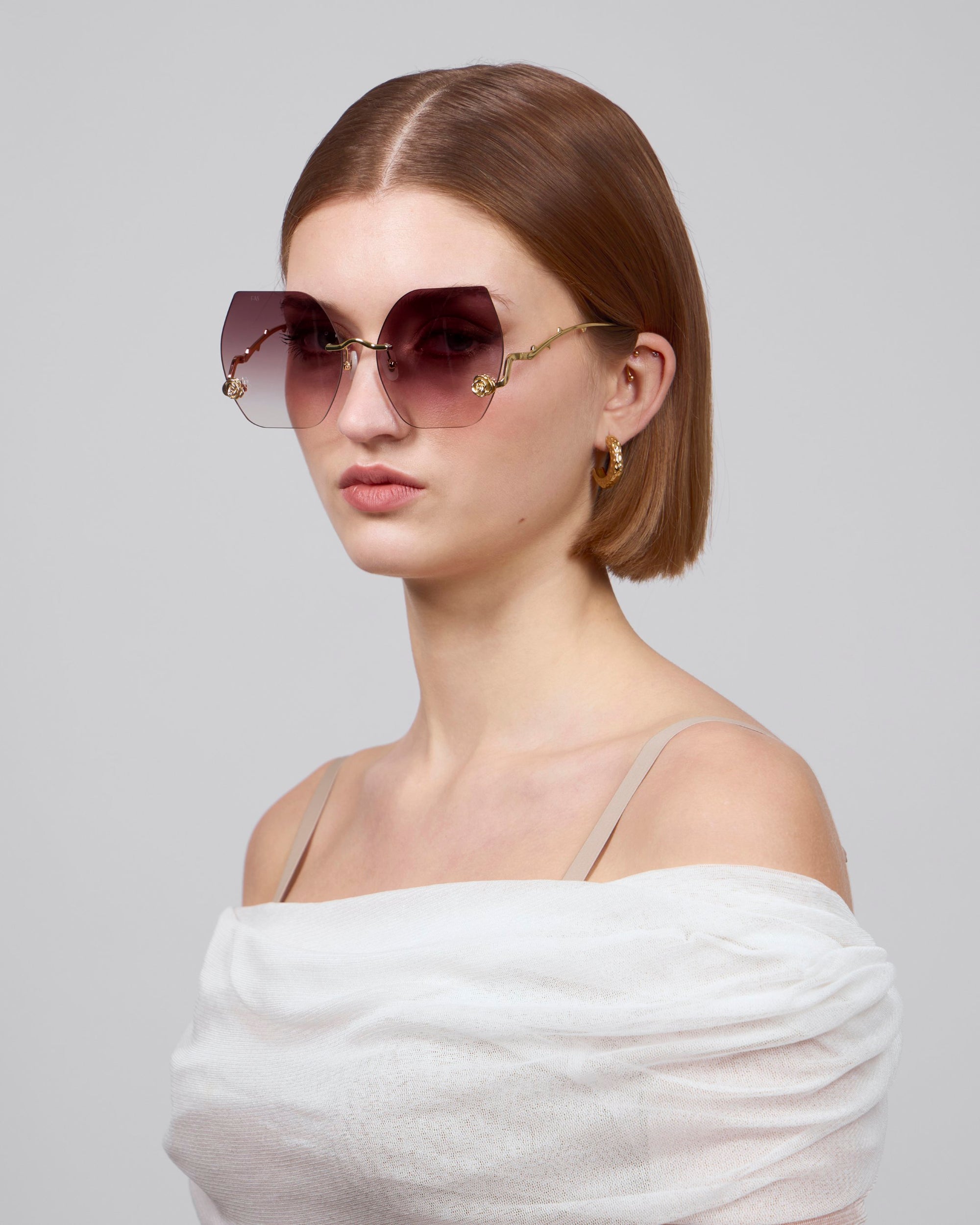 A woman with short brown hair wears retro-inspired La Vie En Rose oversized geometric sunglasses by For Art's Sake®, featuring gold accents and vibrant hoop earrings, dressed in an off-shoulder white top against a plain gray background. The bold eyewear is crafted from biodegradable acetate.