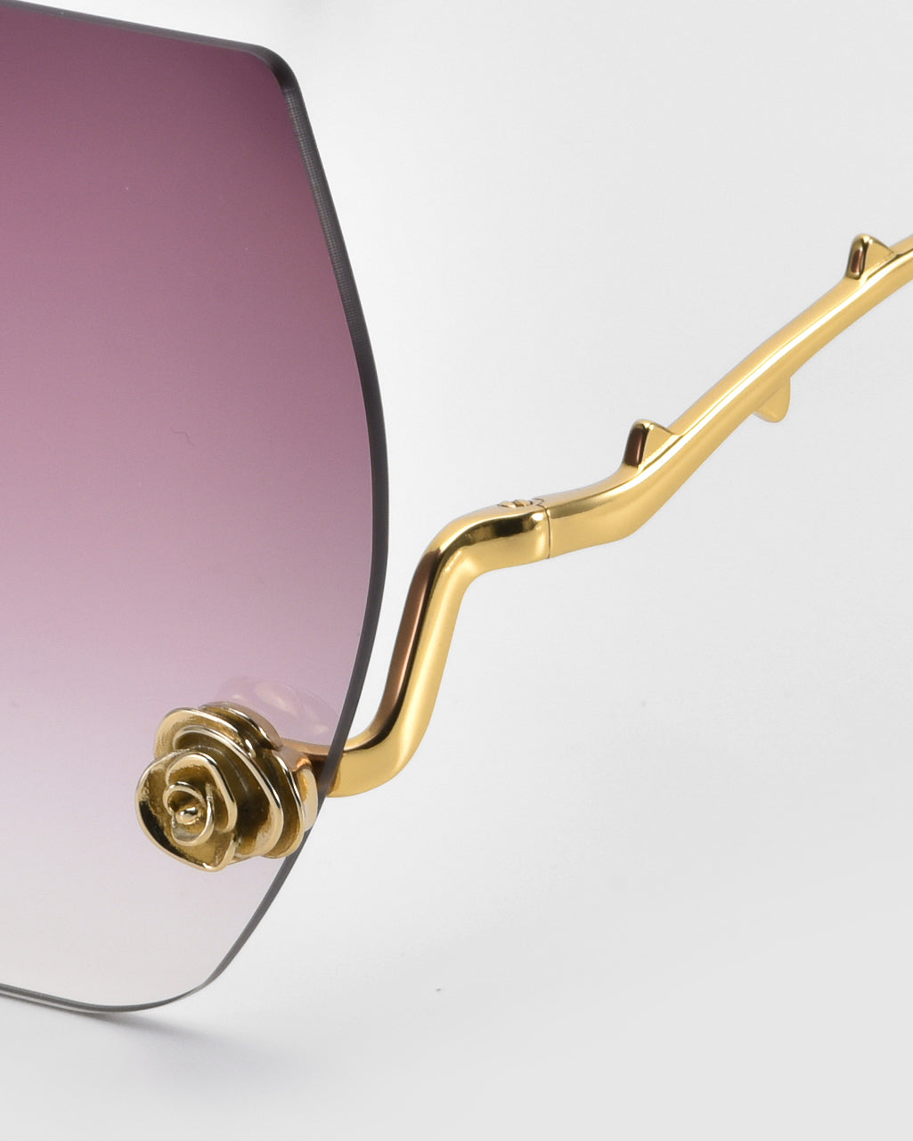 Check out the La Vie En Rose eyeglass frame by For Art's Sake®, part of the Rose Garden Collection. It features oversized frameless lenses with a purple-to-clear gradient, and a gold arm adorned with sculpted rose ornaments at the hinge, leading to a wavy design.