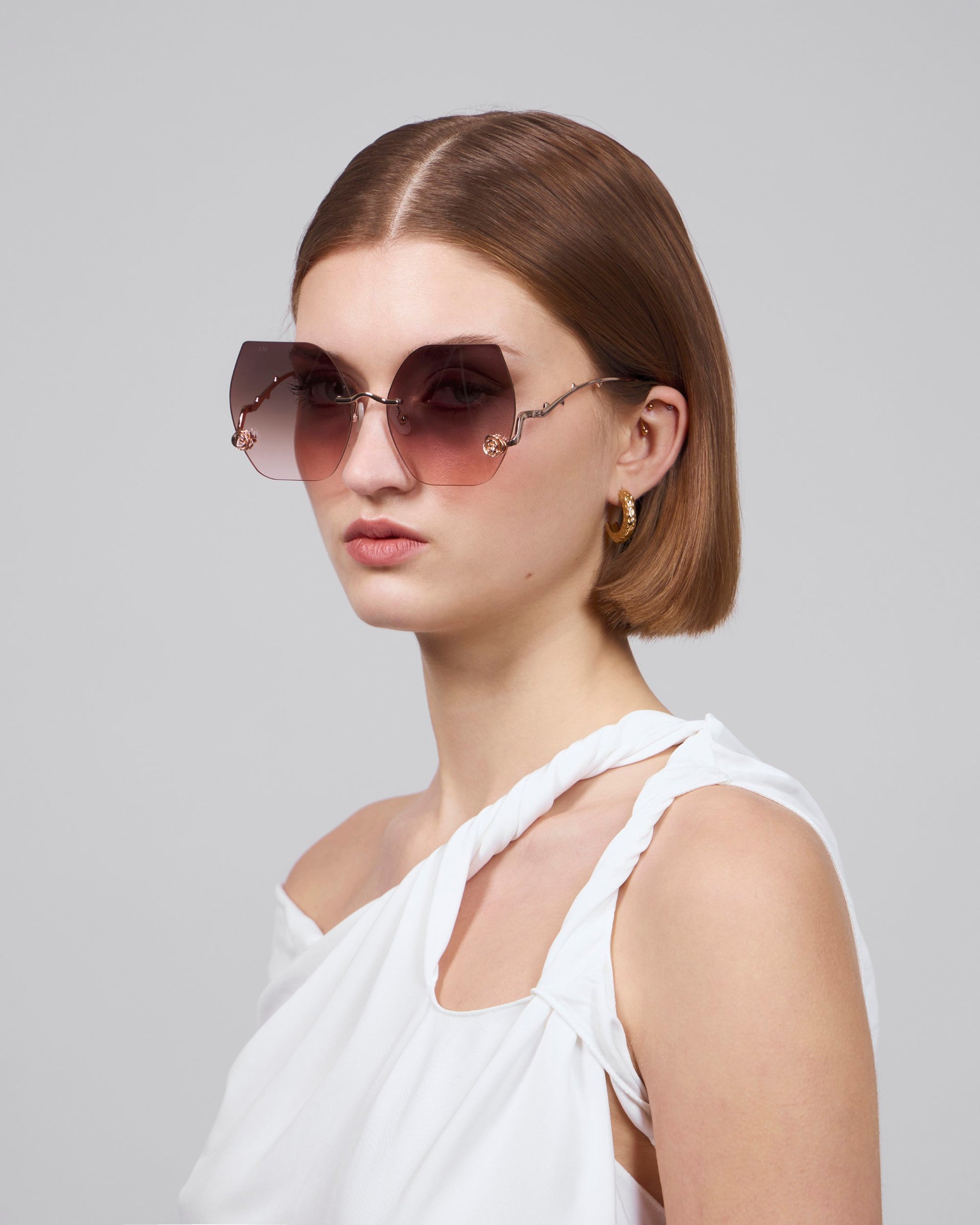 A person with short brown hair wears For Art's Sake® La Vie En Rose oversized square, geometric sunglasses in biodegradable acetate with gradient lenses and gold detailing, paired with a white off-shoulder twisted strap top and small hoop earrings against a plain gray background.