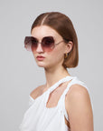 A person with short brown hair wears For Art's Sake® La Vie En Rose oversized square, geometric sunglasses in biodegradable acetate with gradient lenses and gold detailing, paired with a white off-shoulder twisted strap top and small hoop earrings against a plain gray background.