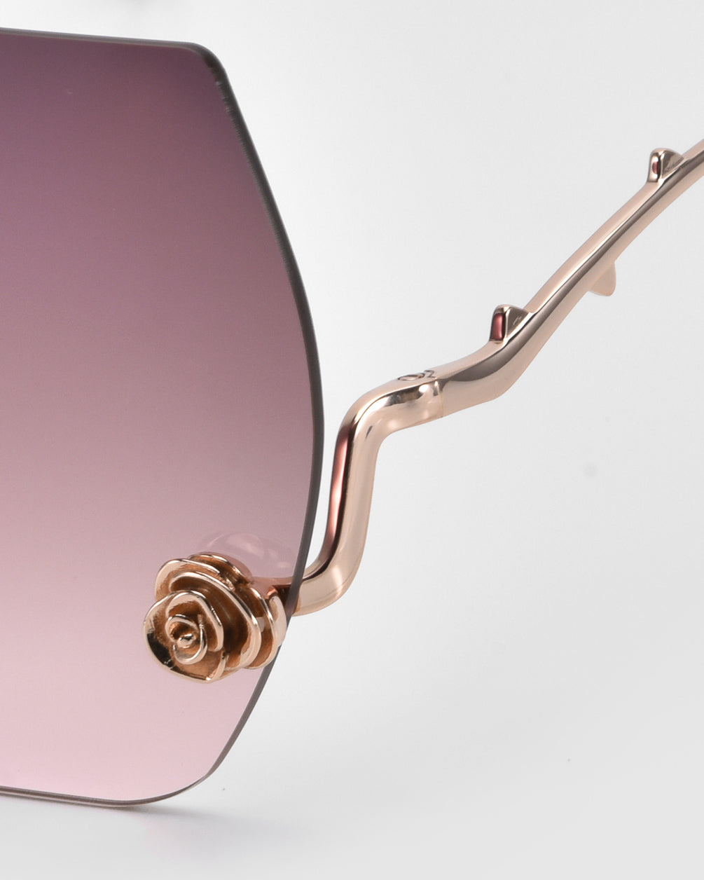 Close-up of La Vie En Rose sunglasses by For Art's Sake®, featuring gradient pink lenses and intricate gold frames. Part of the Rose Garden Collection, they showcase a rose-shaped decoration at the temple for an elegant touch on these stylish oversized frameless shades.