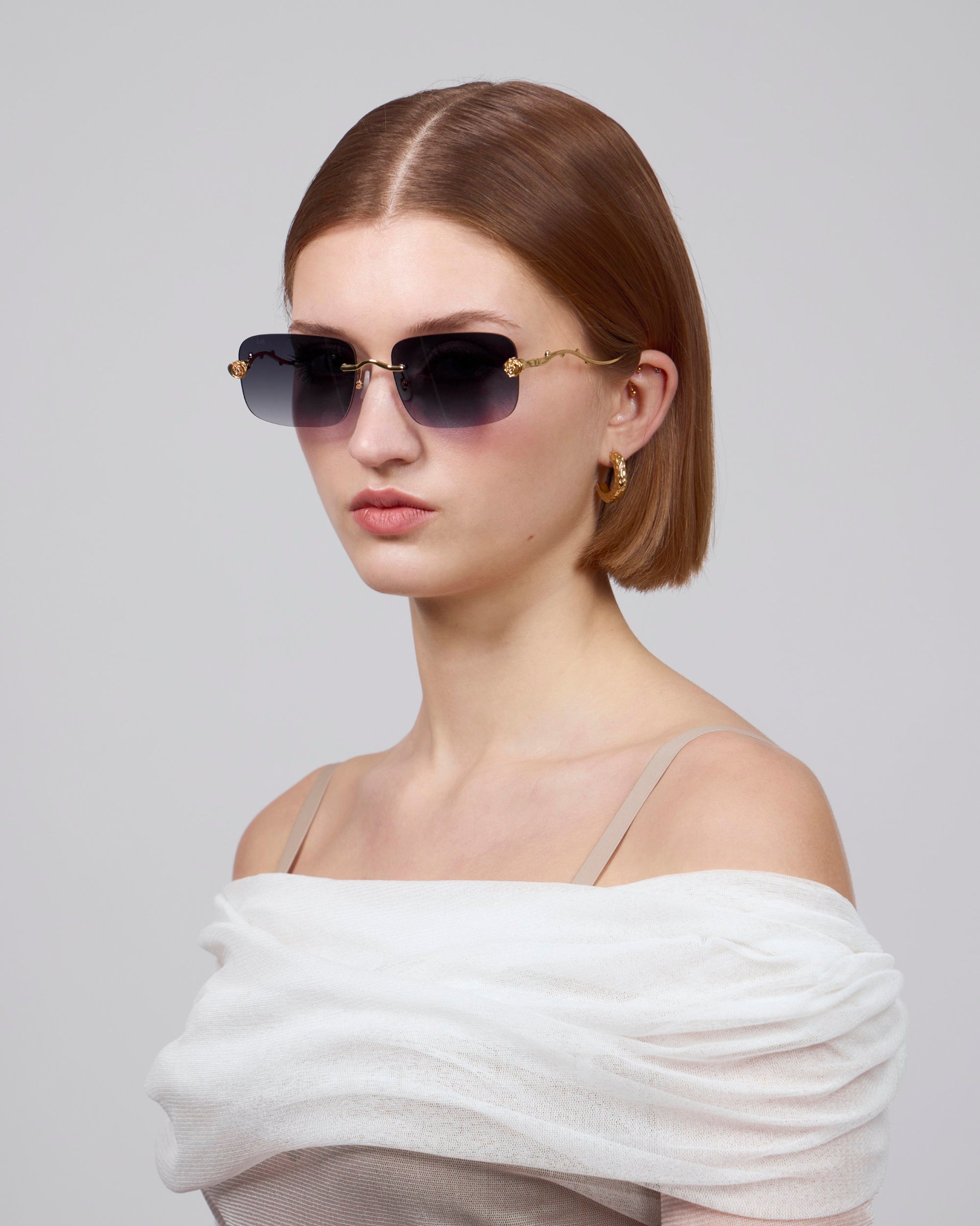 A short-haired woman is styled in gradient oversized frameless lenses from For Art's Sake® Rose Garden Collection and gold hoop earrings, wearing an off-shoulder white top, posing against a plain background.