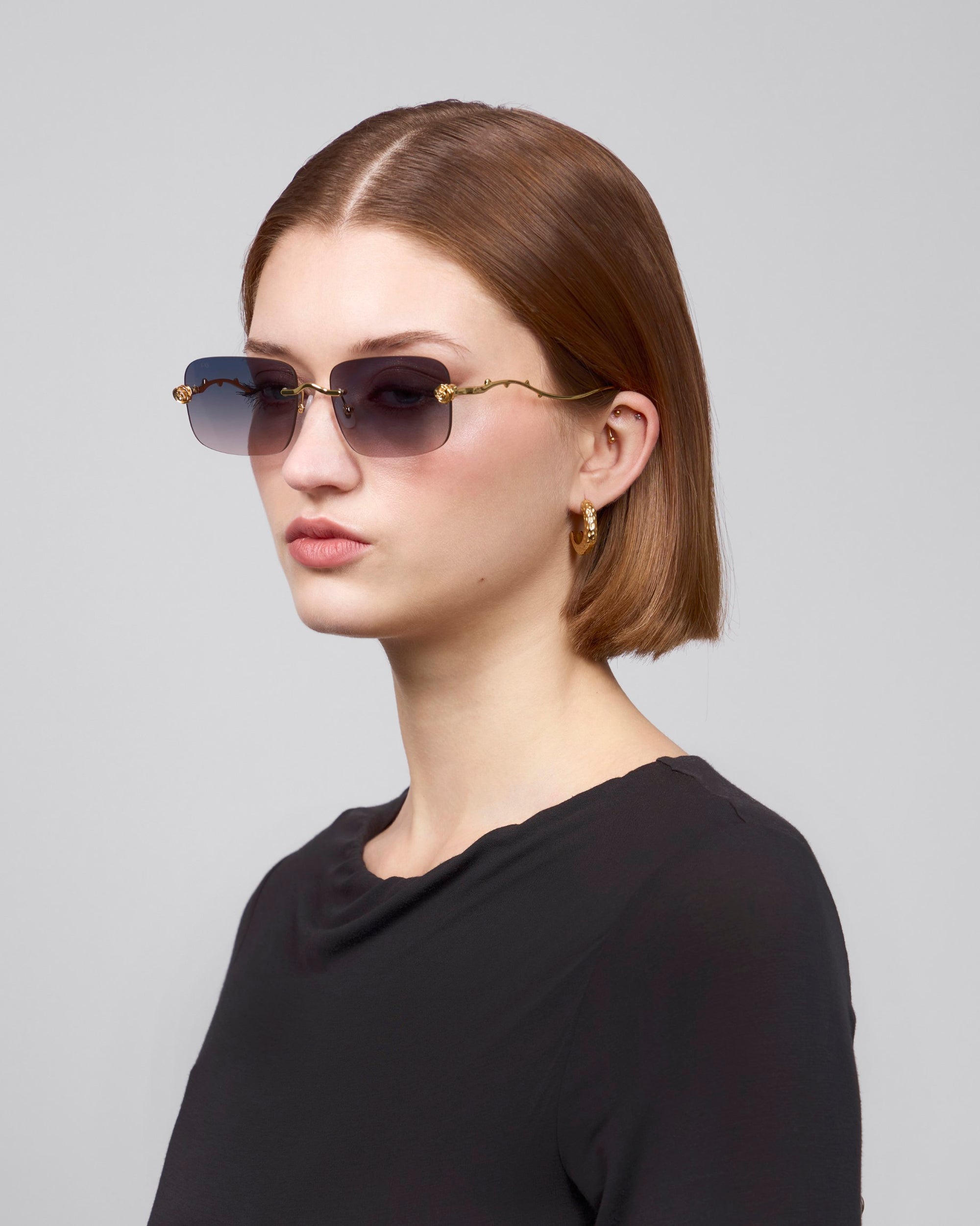 A person with short brown hair, wearing oversized frameless sunglasses from For Art's Sake Rose Garden and gold hoop earrings, is dressed in a black top. They are looking to the side against a plain light gray background.