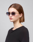 A person with short brown hair, wearing oversized frameless sunglasses from For Art's Sake Rose Garden and gold hoop earrings, is dressed in a black top. They are looking to the side against a plain light gray background.