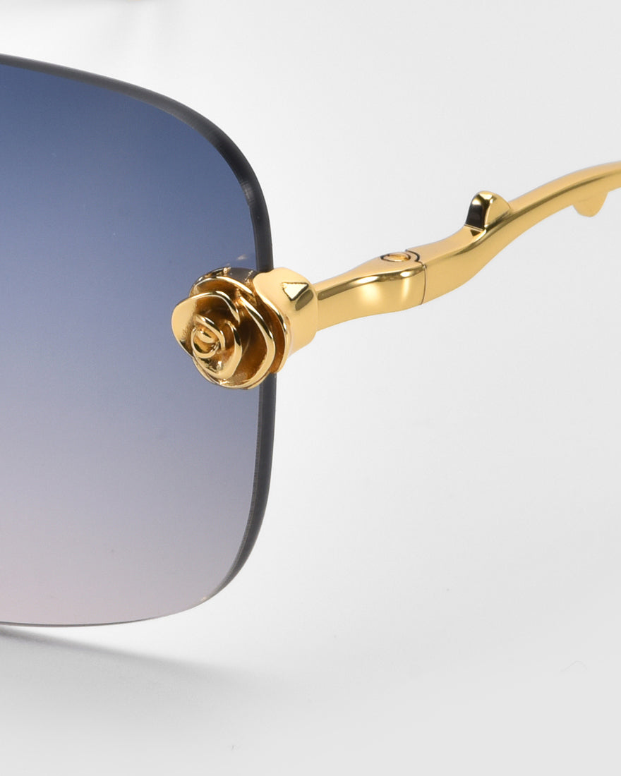 Close-up of For Art's Sake® Rose Garden sunglasses showcasing gradient lenses. The frame has sculpted rose ornaments and stylized branch designs on the temple. A lens corner is visible against a light gray background.