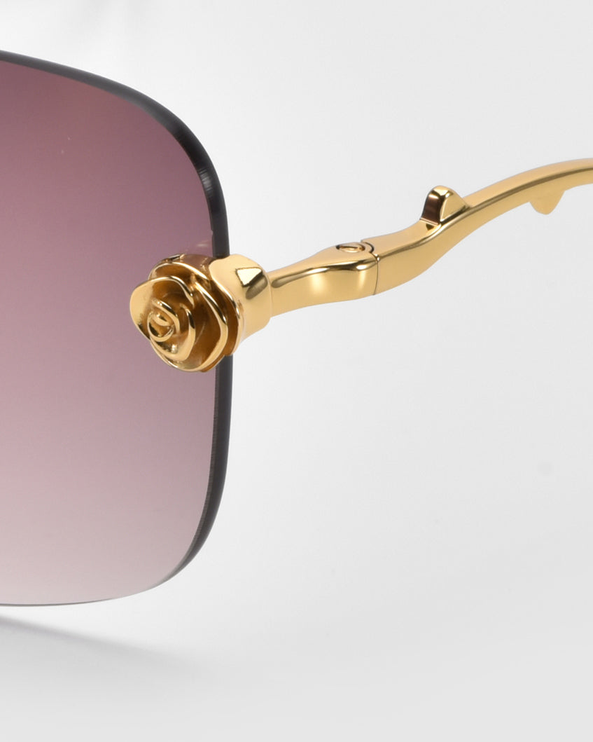 A close-up of For Art's Sake® Rose Garden sunglasses showcases square, gradient-tinted lenses, and gold temples with sculpted rose ornaments by the hinge. The captivating dark-to-light gradient of the lenses contrasts beautifully with a plain, light-colored background.