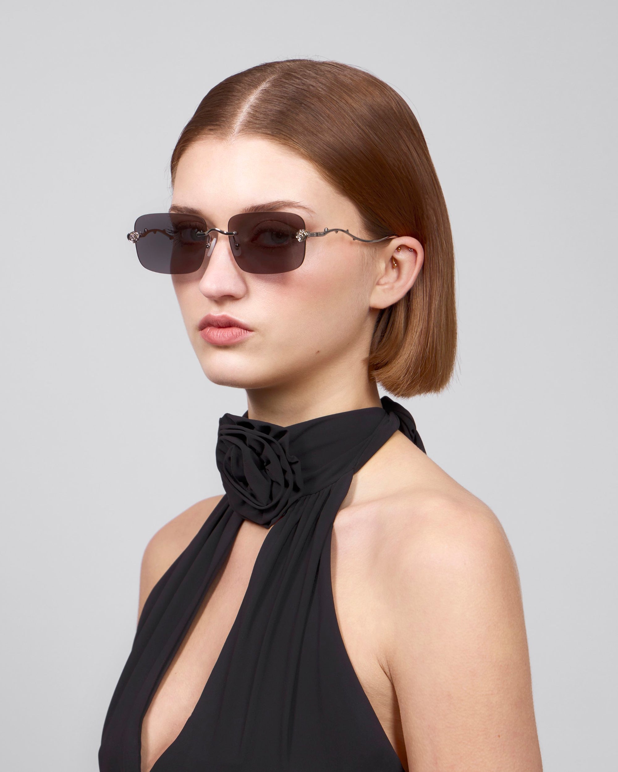 A person with straight, shoulder-length brown hair wears oversized frameless sunglasses from For Art's Sake® Rose Garden collection and a black halter top with floral detail at the neck. They pose against a plain gray background with a neutral expression.