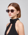 A person with straight, shoulder-length brown hair wears oversized frameless sunglasses from For Art's Sake® Rose Garden collection and a black halter top with floral detail at the neck. They pose against a plain gray background with a neutral expression.