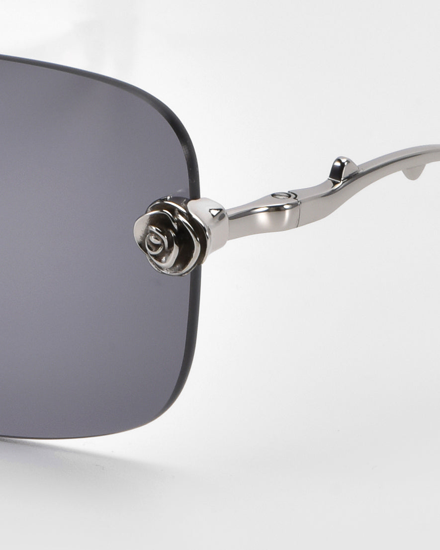 A close-up of stylish For Art's Sake® Rose Garden sunglasses with dark lenses and a silver metal frame featuring sculpted rose ornaments on the temple. The background is plain white.