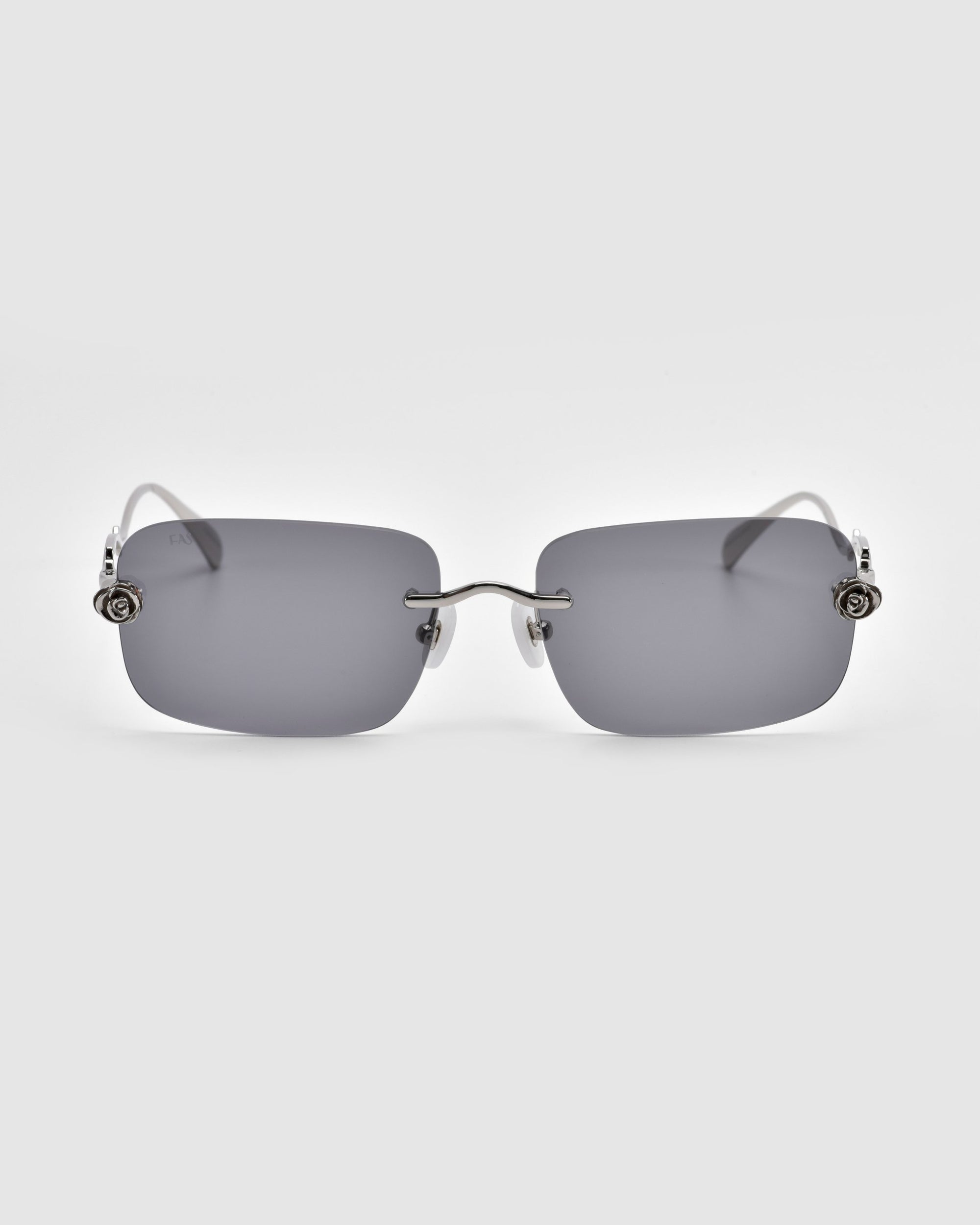 These Rose Garden rimless sunglasses from For Art's Sake® feature rectangular gray lenses and thin silver arms adorned with decorative metal details near the hinges, reminiscent of sculpted rose ornaments.