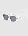 The stylish Rose Garden sunglasses by For Art's Sake® boast oversized frameless lenses with rectangular gray shades and delicate metal arms, set against a light gray backdrop.