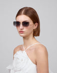 A short-haired brunette with an artistic flair, wears For Art's Sake® Vermilion rectangular rimless sunglasses and gold hoop earrings. She is in a white top featuring a large fabric rose. A plain gray background completes the look.