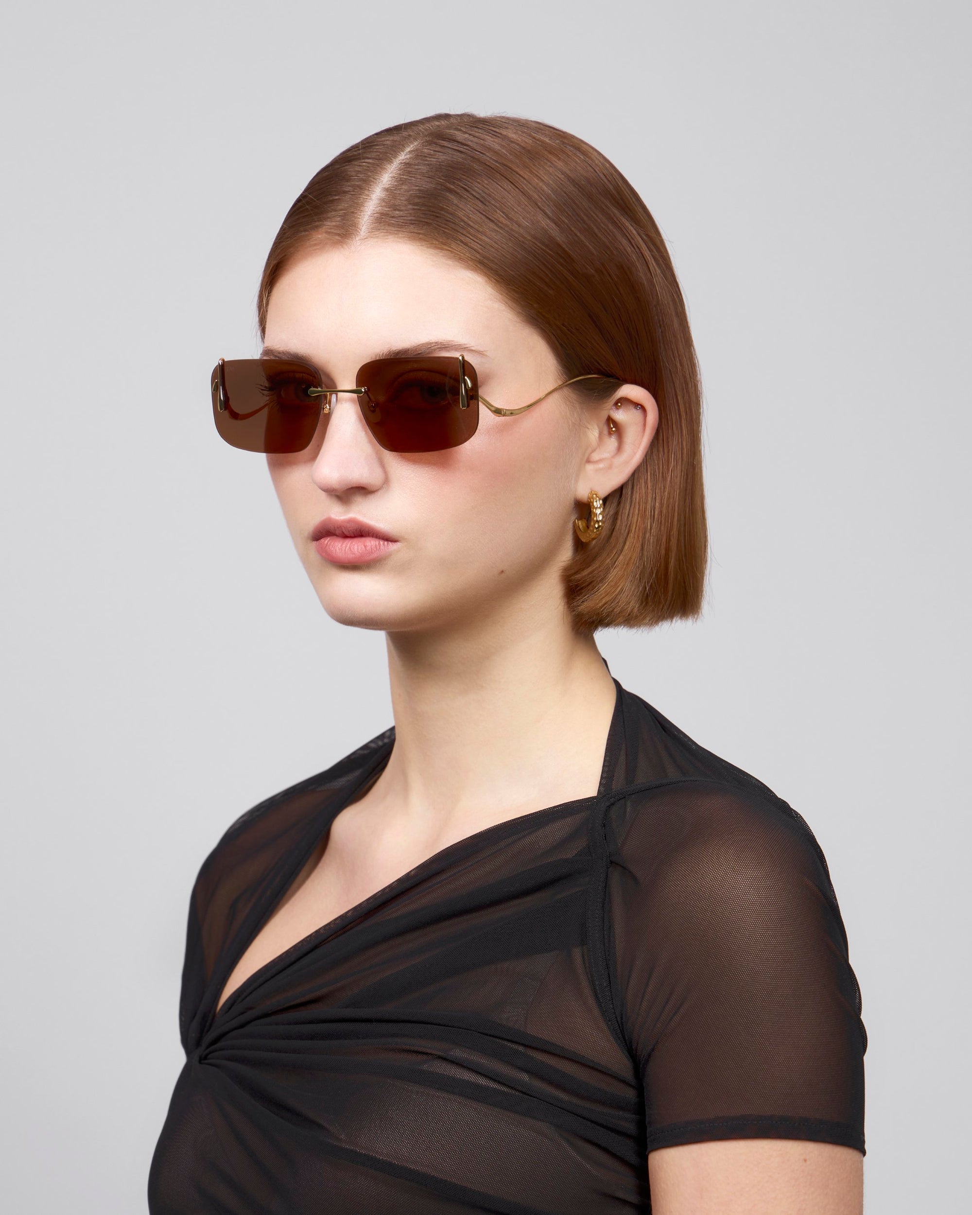 A short-haired person wearing Vermilion rectangular sunglasses by For Art's Sake®, gold hoop earrings, and a black sheer top poses against a gray background, exuding artistic flair.