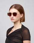 A short-haired person wearing Vermilion rectangular sunglasses by For Art's Sake®, gold hoop earrings, and a black sheer top poses against a gray background, exuding artistic flair.