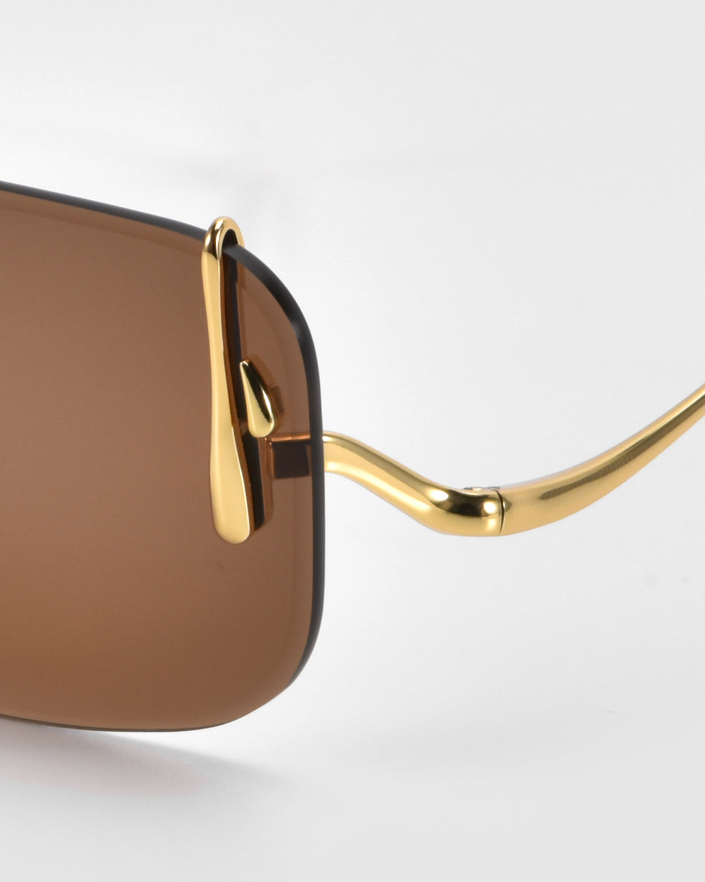 A close-up of For Art's Sake® Vermilion sunglasses shows a rimless silhouette with rectangular brown lenses and a unique gold frame. Elegantly sculptural temples give a sophisticated touch against a plain white background.