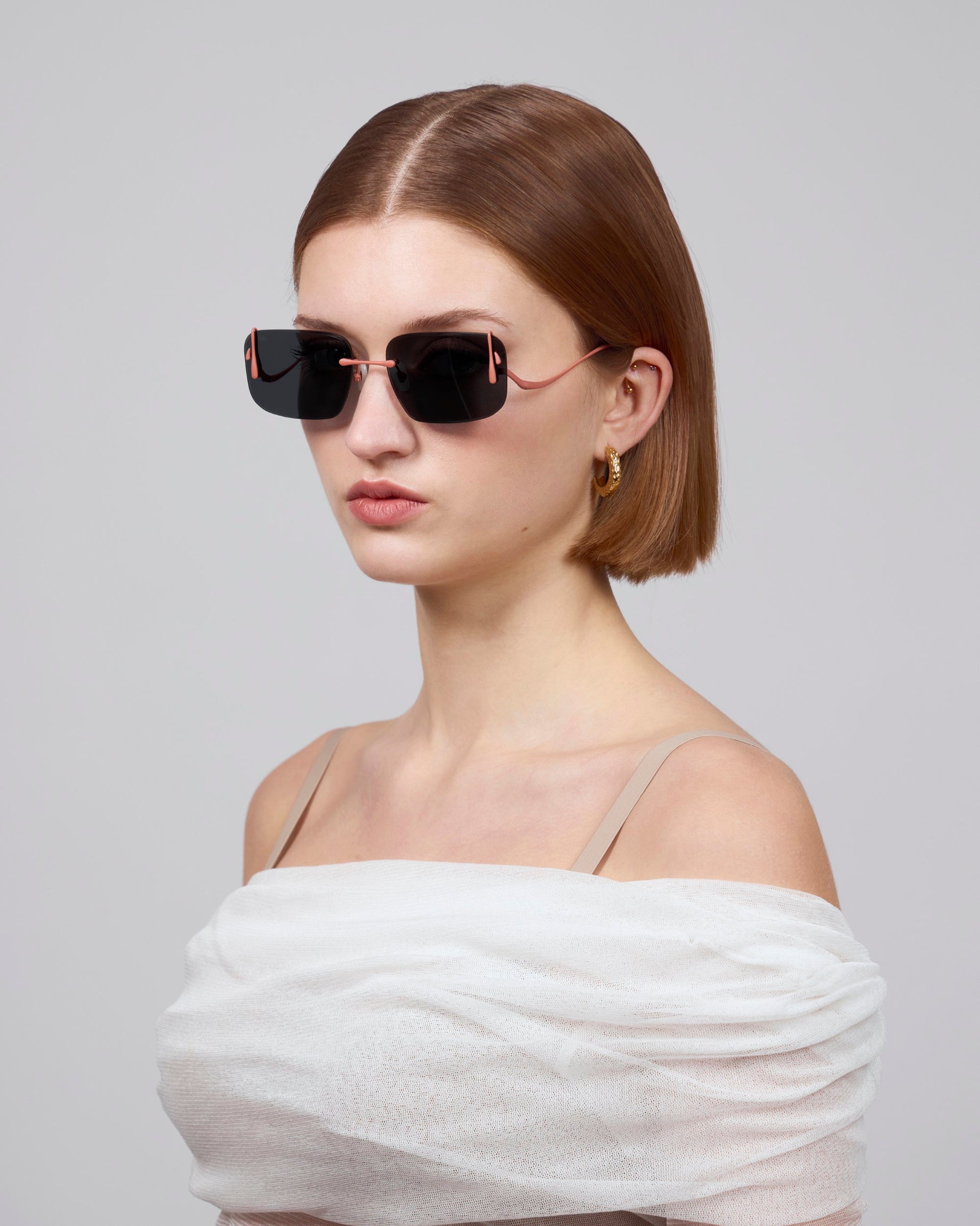 An artistic individual with short brown hair wears For Art's Sake® black sunglasses and gold hoop earrings, styled in a white off-the-shoulder Vermilion top against a light gray background.