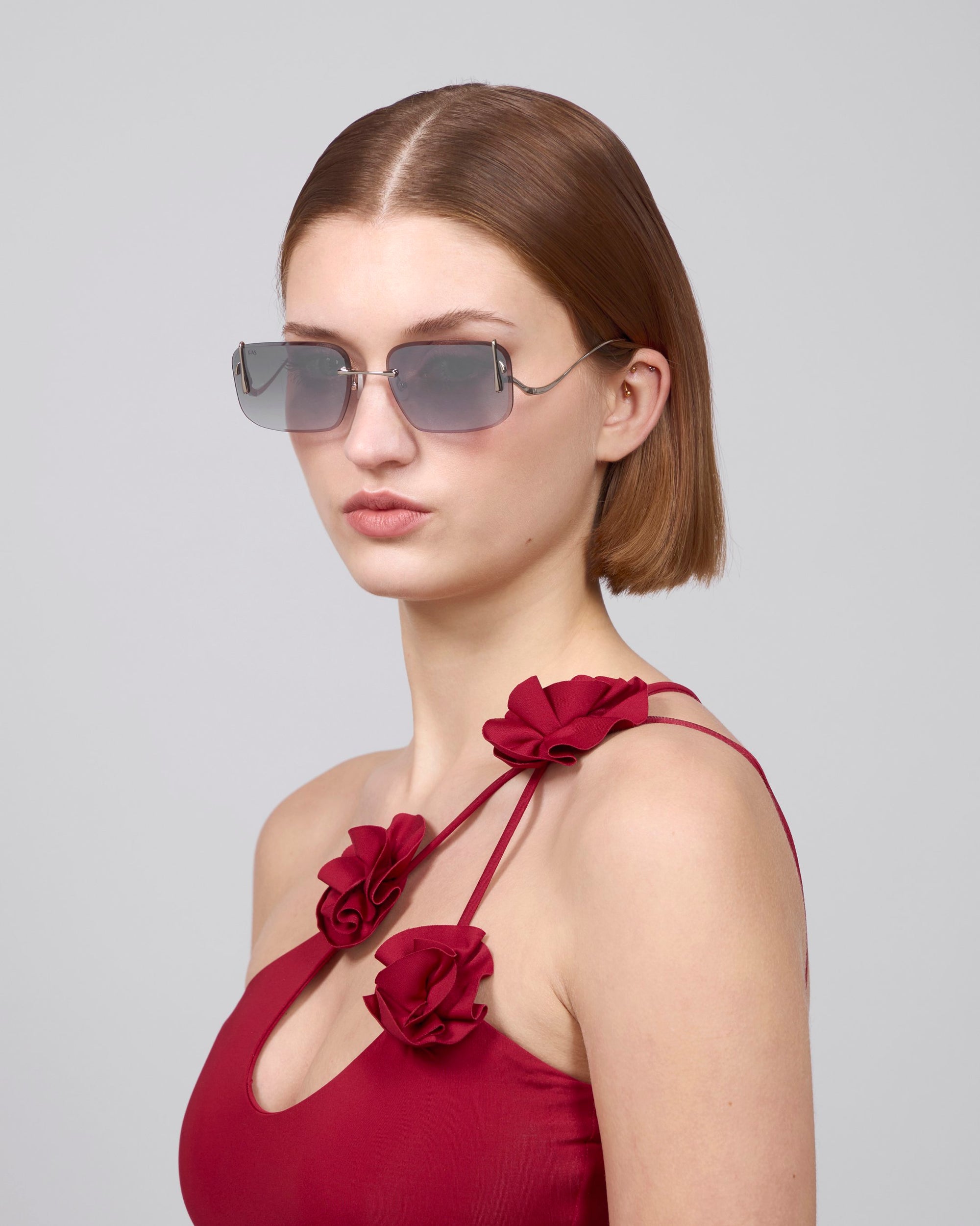 A person with straight, short hair and oversized For Art's Sake® sunglasses with sculptural temples wears the Vermilion red dress featuring floral embellishments on the shoulder straps, posing against a plain backdrop.