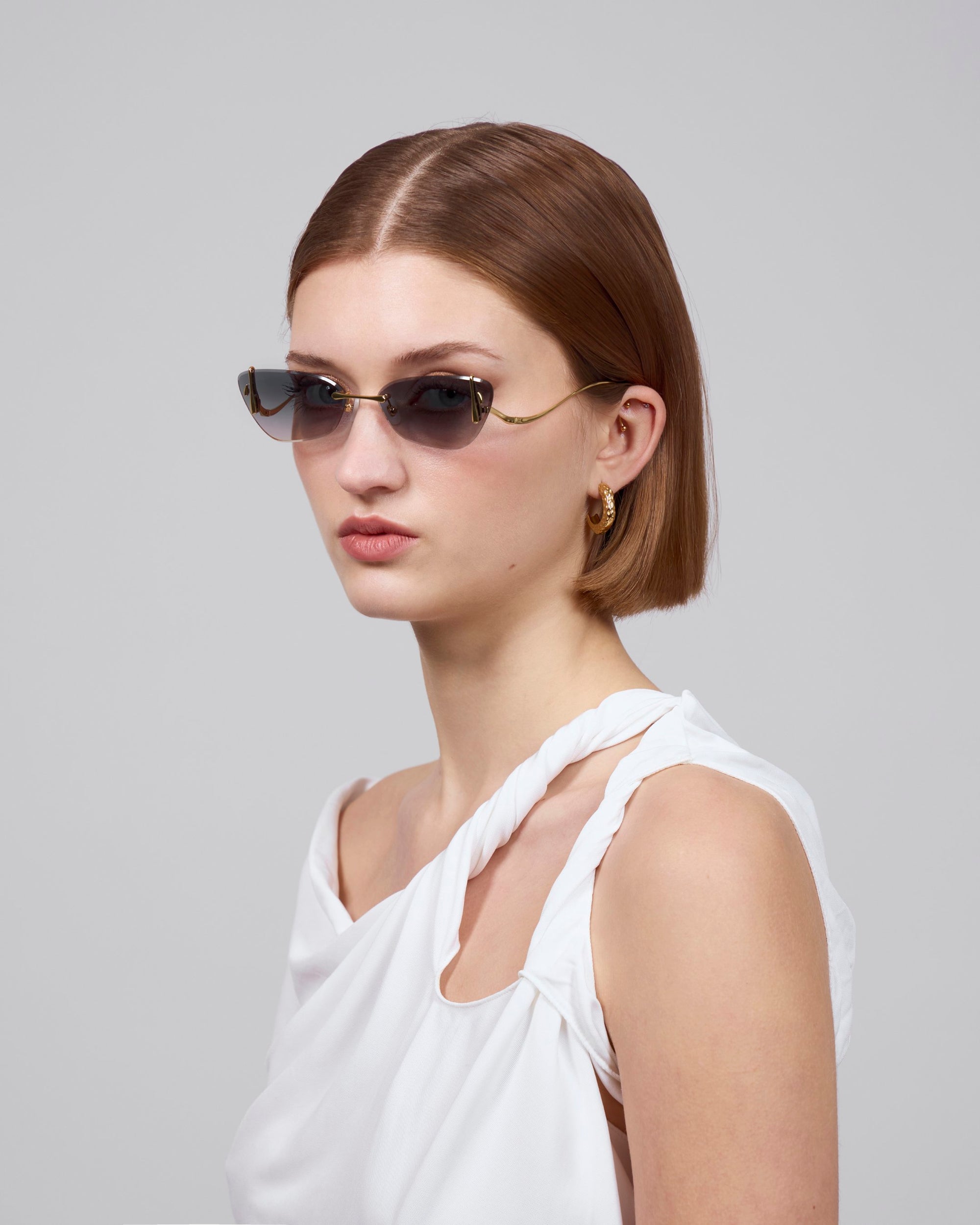 A person with short brown hair, wearing thin-framed sunglasses and a white, sleeveless twisted-strap top from For Art's Sake® Rose Garden Collection, looks to the side against a plain background. Sculpted rose ornaments complete their look with enchanting duality enhanced by gold hoop earrings.