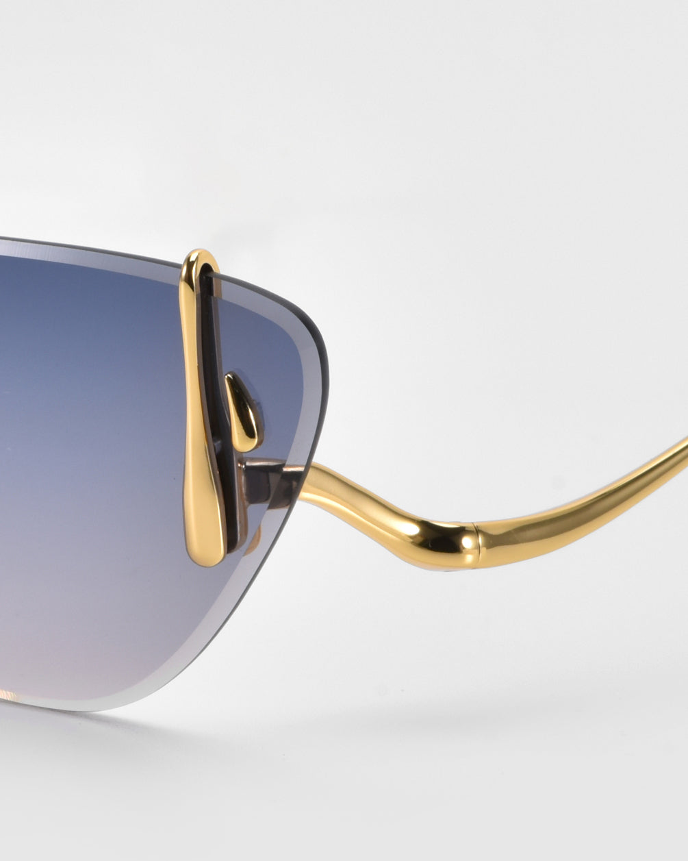 A close-up showcases For Art's Sake® Crimson sunglasses against a light gray background. These stylish, lightweight glasses feature gradient lenses transitioning from blue to clear in a sleek, modern rimless design with glossy gold accents.