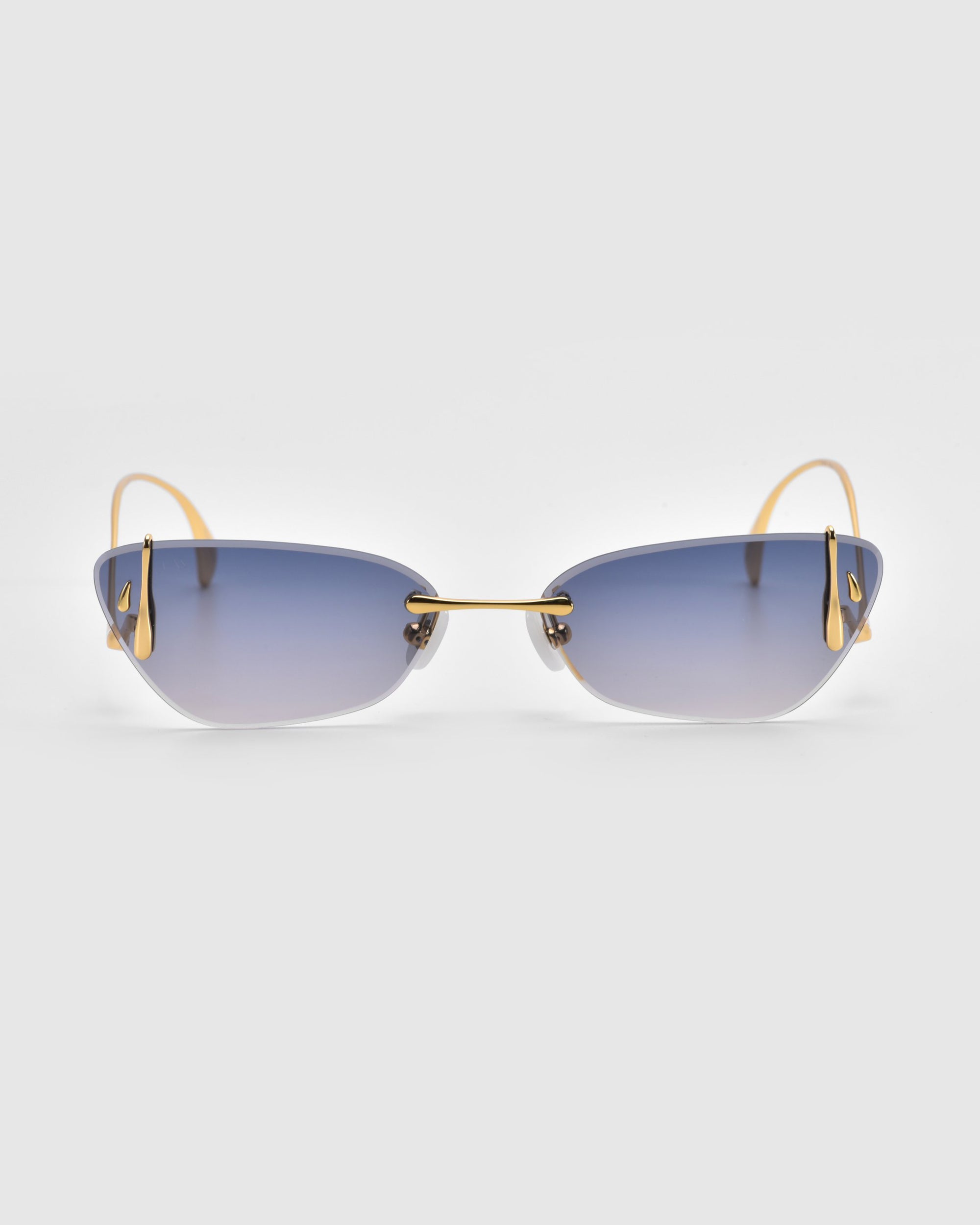 Crimson sunglasses from For Art's Sake® feature elegant gradient blue lenses, gold wireframe arms, a sculpted rose ornament on the gold nose bridge, and stylishly angled frameless edges. Seen from the front against a light gray backdrop. Part of the Rose Garden Collection.