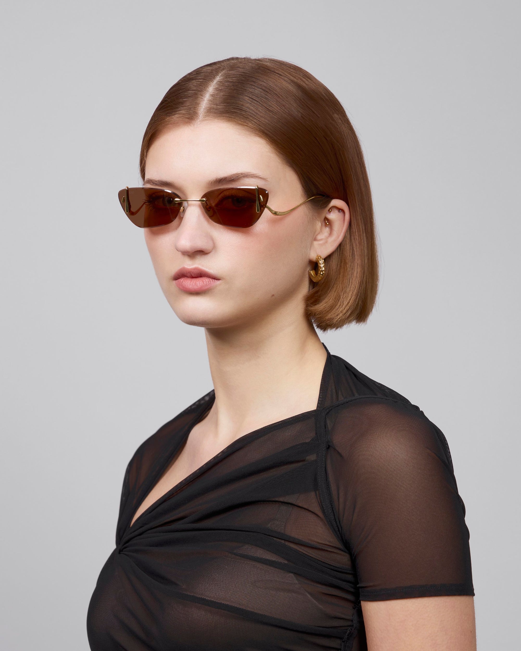 A person with shoulder-length brown hair wears angular brown sunglasses and gold hoop earrings from For Art’s Sake®&#39;s Rose Garden Collection. Their neutral expression complements a sheer black top, embodying an enchanting duality against a plain light gray background.