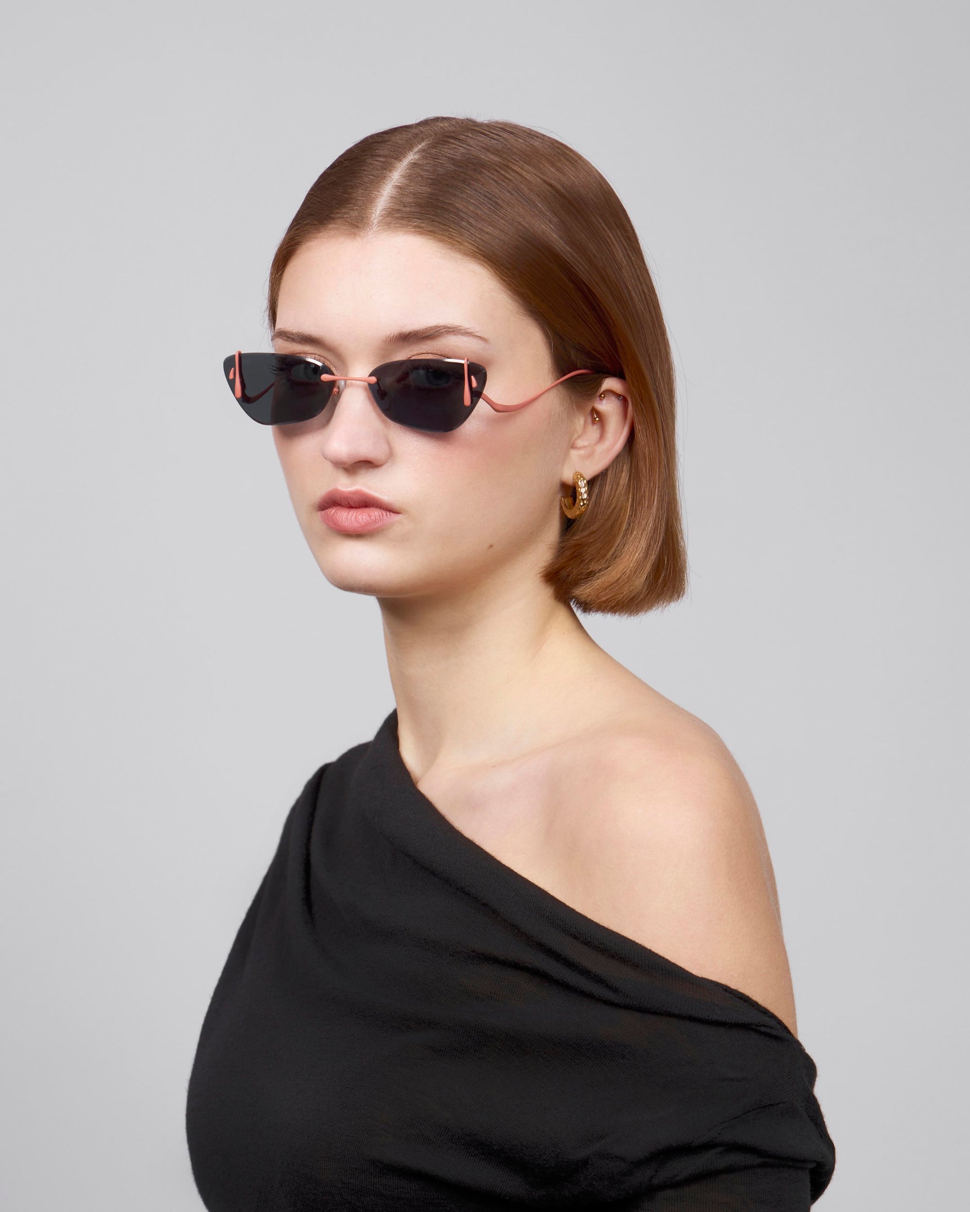 A person with straight, shoulder-length brown hair wears dark sunglasses and the &quot;Crimson&quot; black, off-the-shoulder top from For Art&#39;s Sake® on a plain gray background, highlighting the enchanting duality of sculpted rose ornaments subtly accentuating their look.