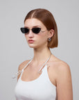 A person with short brown hair, in dark sunglasses, wears 'Crimson' from For Art's Sake® and silver hoop earrings. A white neck strap completes the look against a plain gray backdrop, highlighting the elegance of this ensemble.