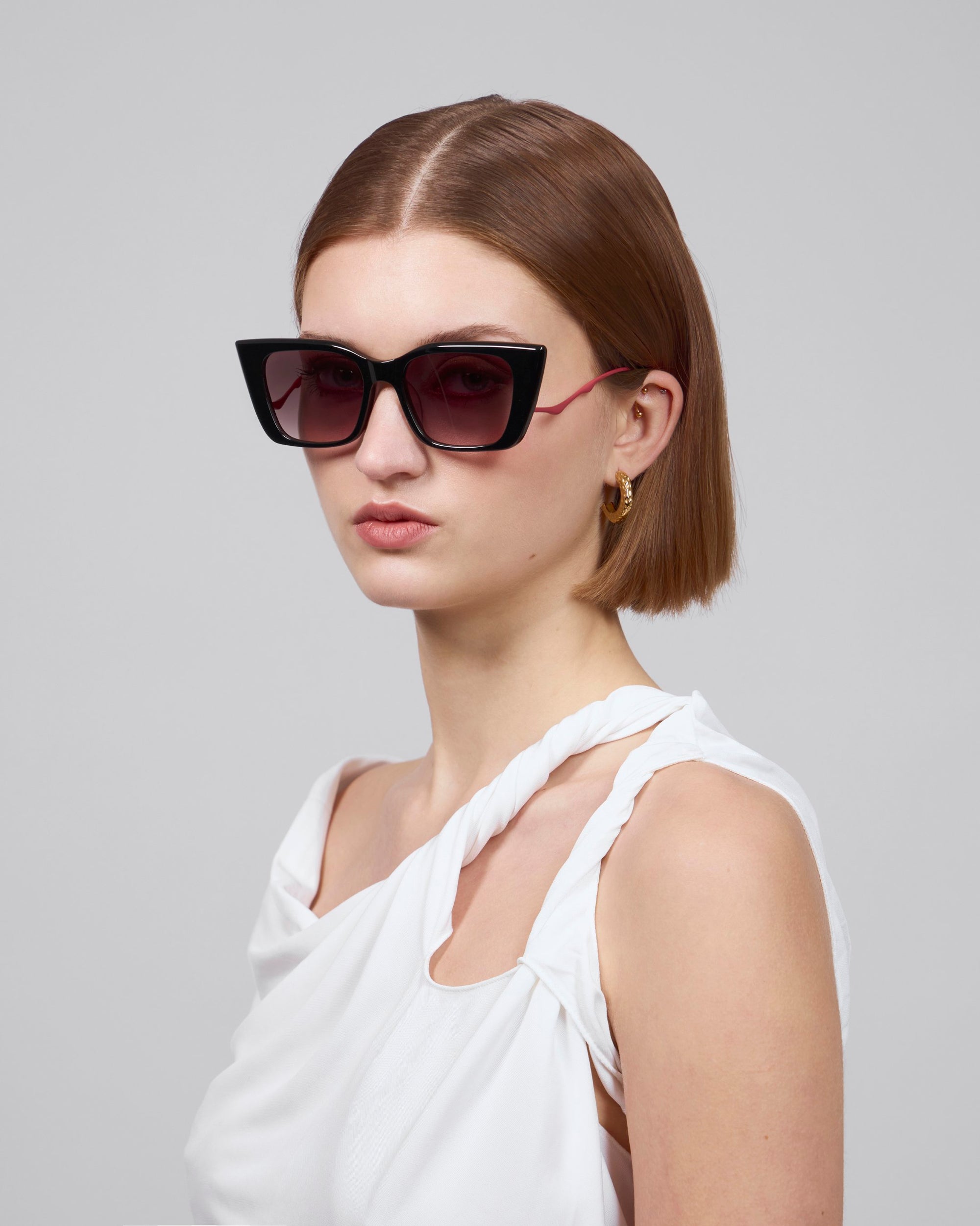 A person with short, straight brown hair wears oversized "Dusk" sunglasses by For Art's Sake® with an acetate front frame. They're dressed in a sleeveless white top, gold hoop earrings, and posed against a plain gray background.