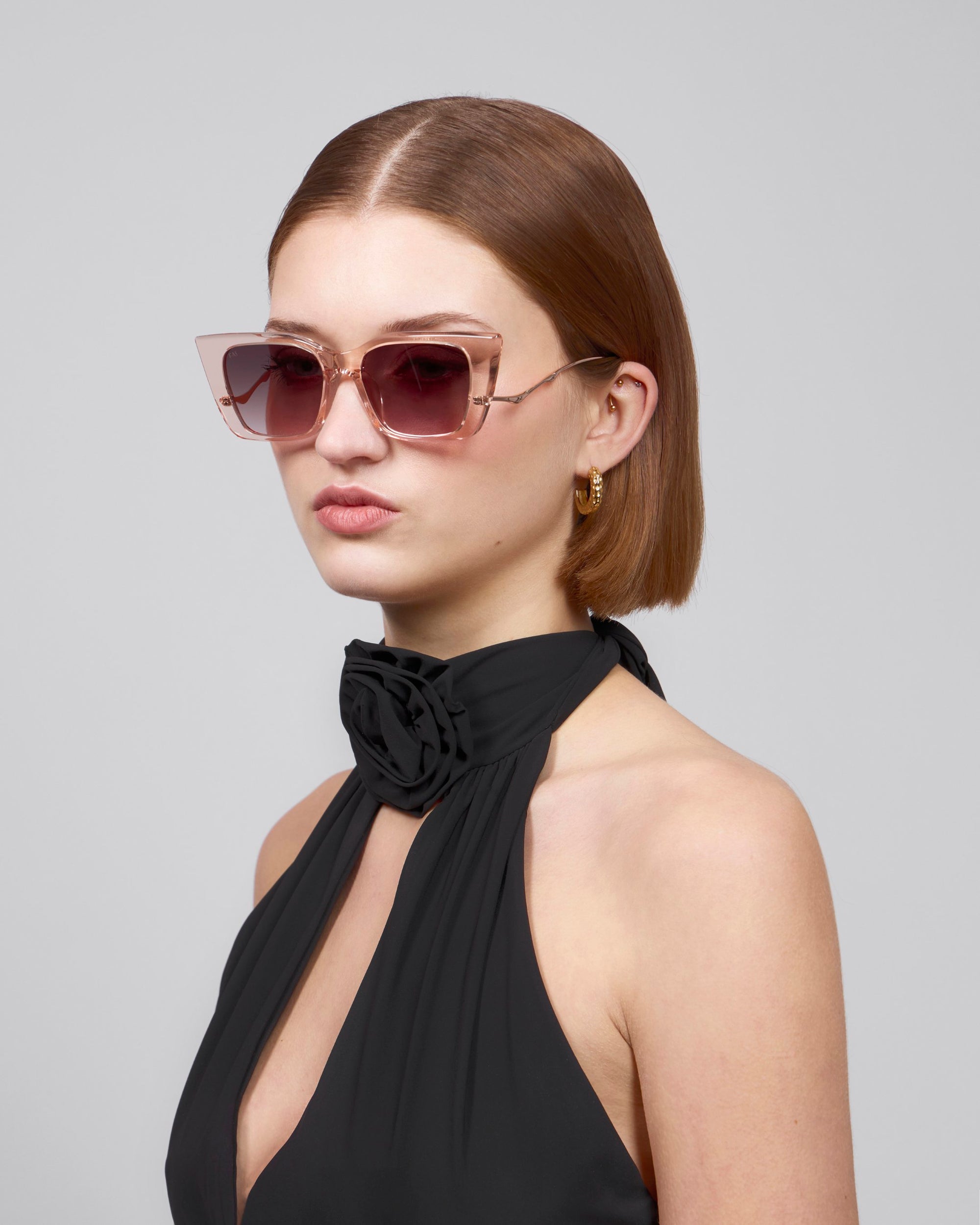 In a plain grey setting, a short-haired woman exudes sophistication in For Art's Sake® Dusk pink-tinted, chunky cat-eye sunglasses and a black halter dress with a floral accent, gazing to the side.